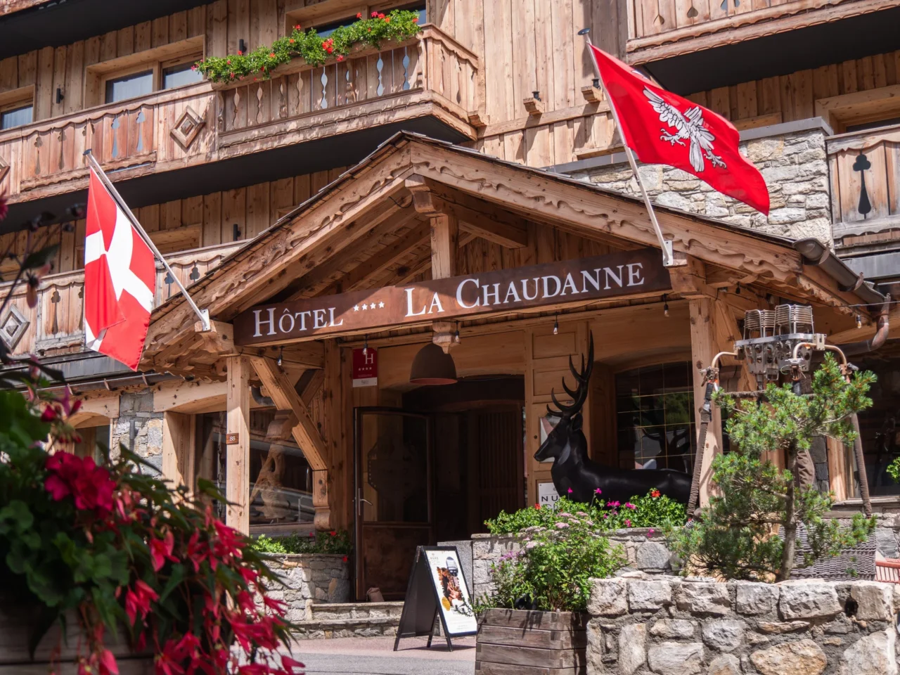 Entrée de l’Hôtel La Chaudanne sous un porche en bois, avec balcons fleuris et façade en pierre et bois.