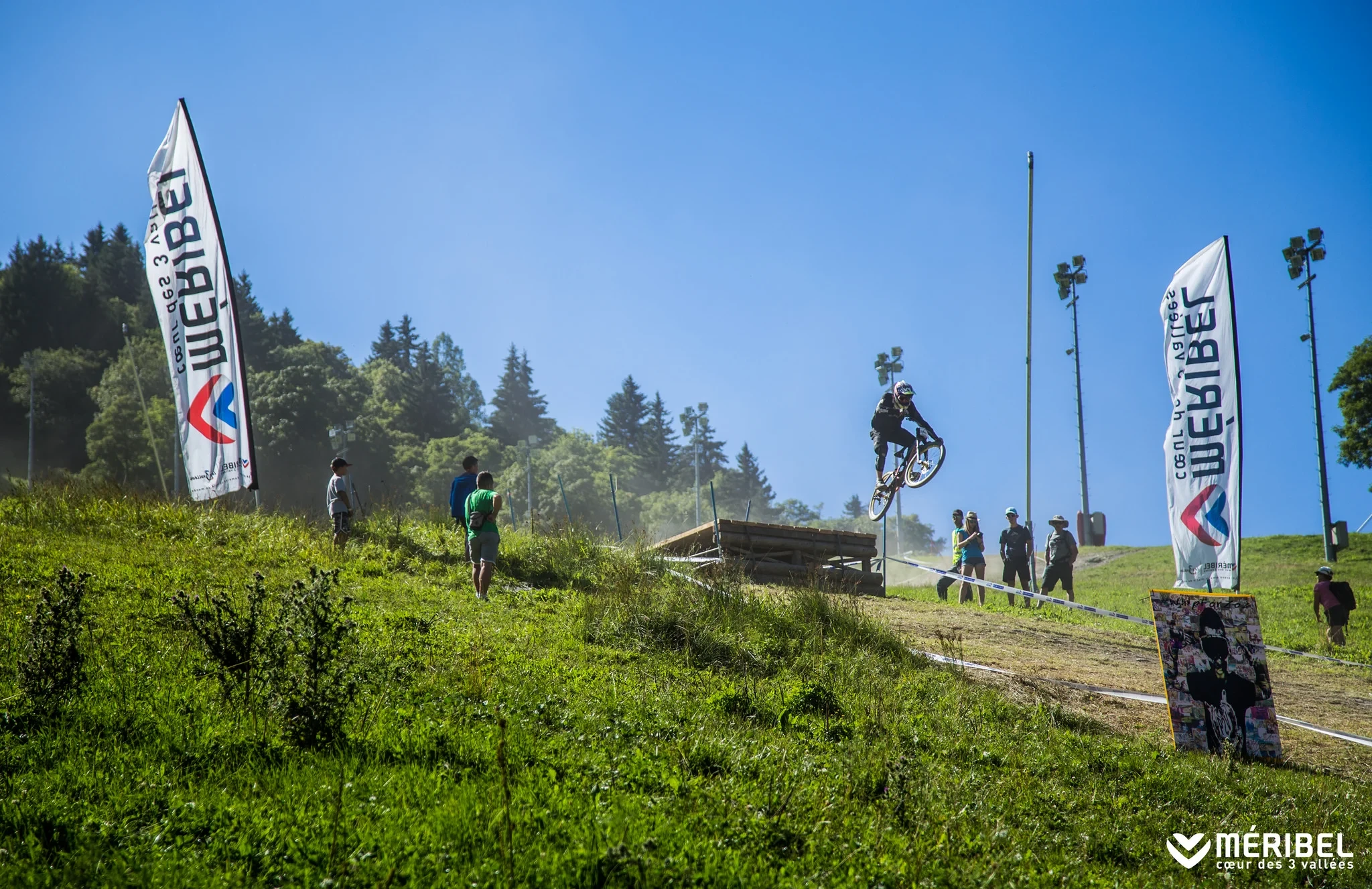 Un vététiste effectue un saut au-dessus d’une rampe en bois, entre deux drapeaux de Méribel.