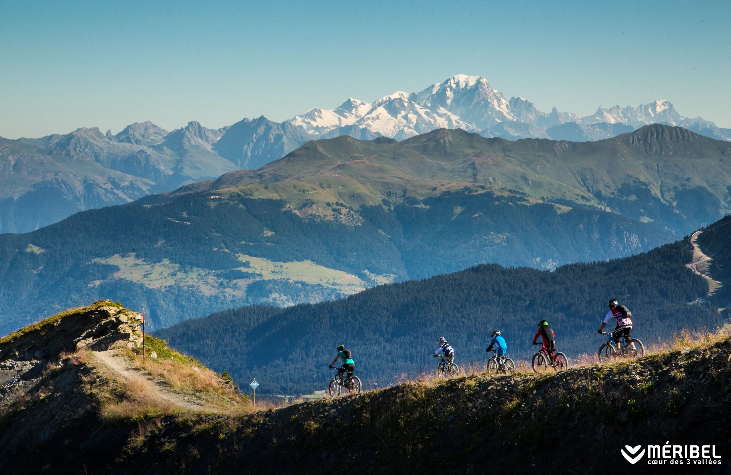 Cinq vététistes sur un sentier de montagne à Méribel en été, face aux sommets enneigés.