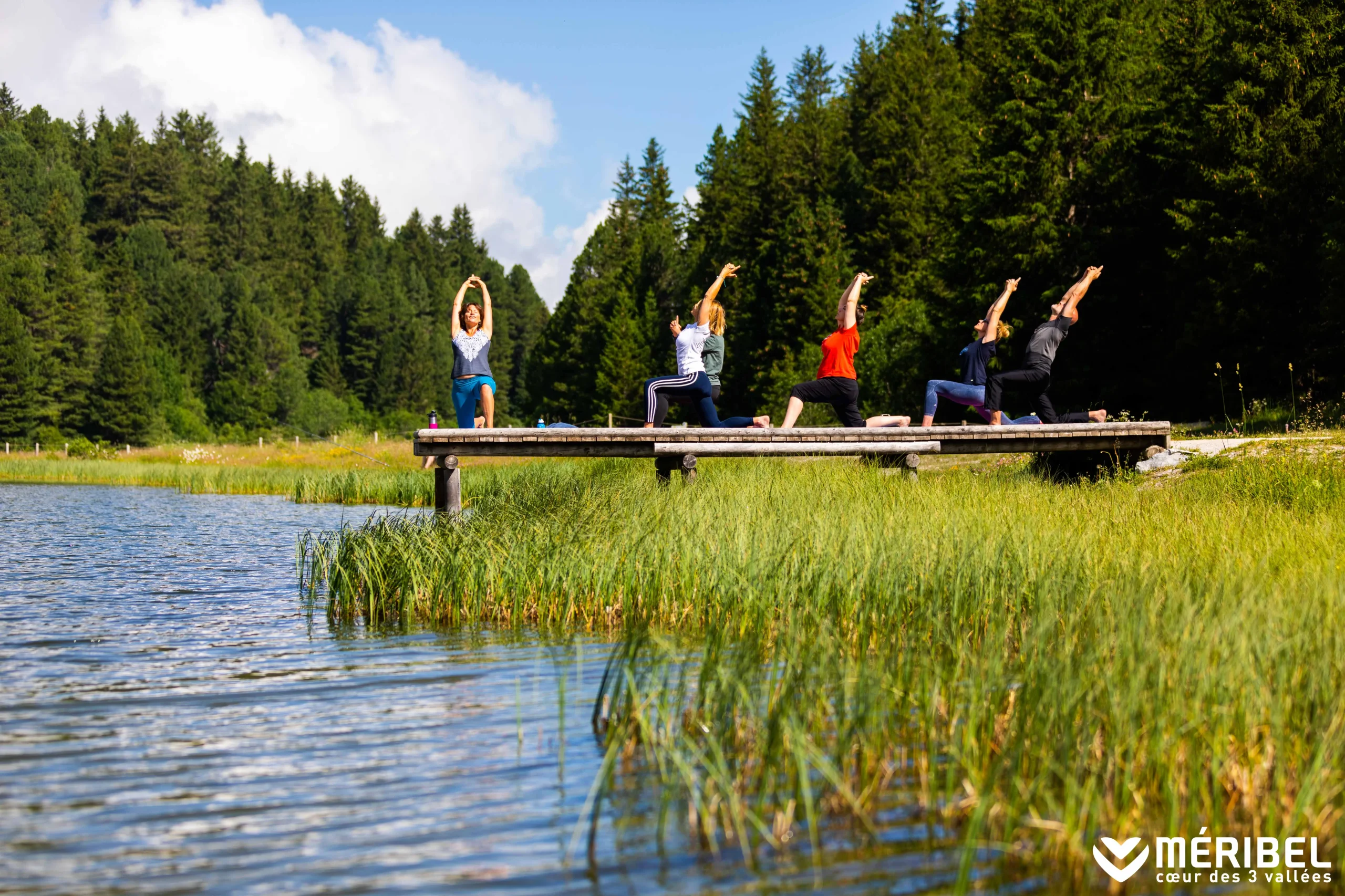 Séance de yoga sur une plateforme en bois au bord d’un lac à Méribel, devant une forêt de conifères.