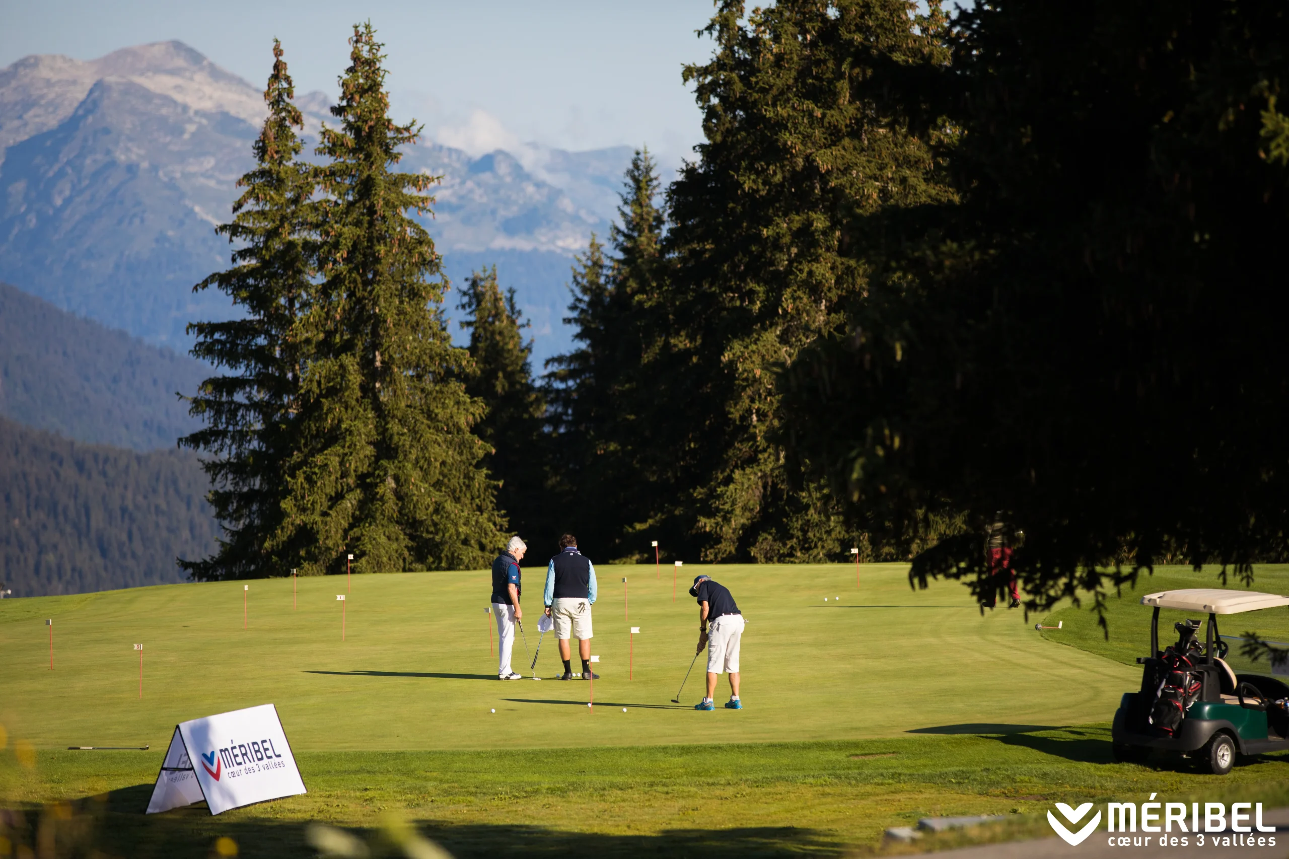 Des golfeurs s’entraînent au putting sur un green de Méribel entouré de conifères et de montagnes.