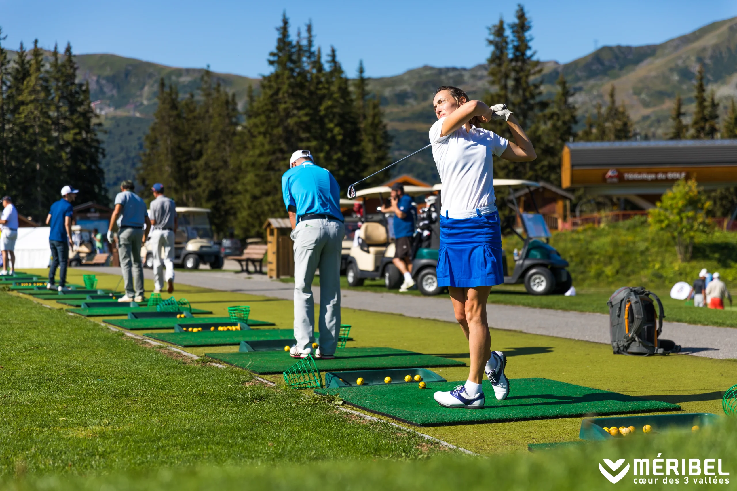 Golfeuse terminant son swing sur un tapis d’entraînement, avec les montagnes en arrière-plan.