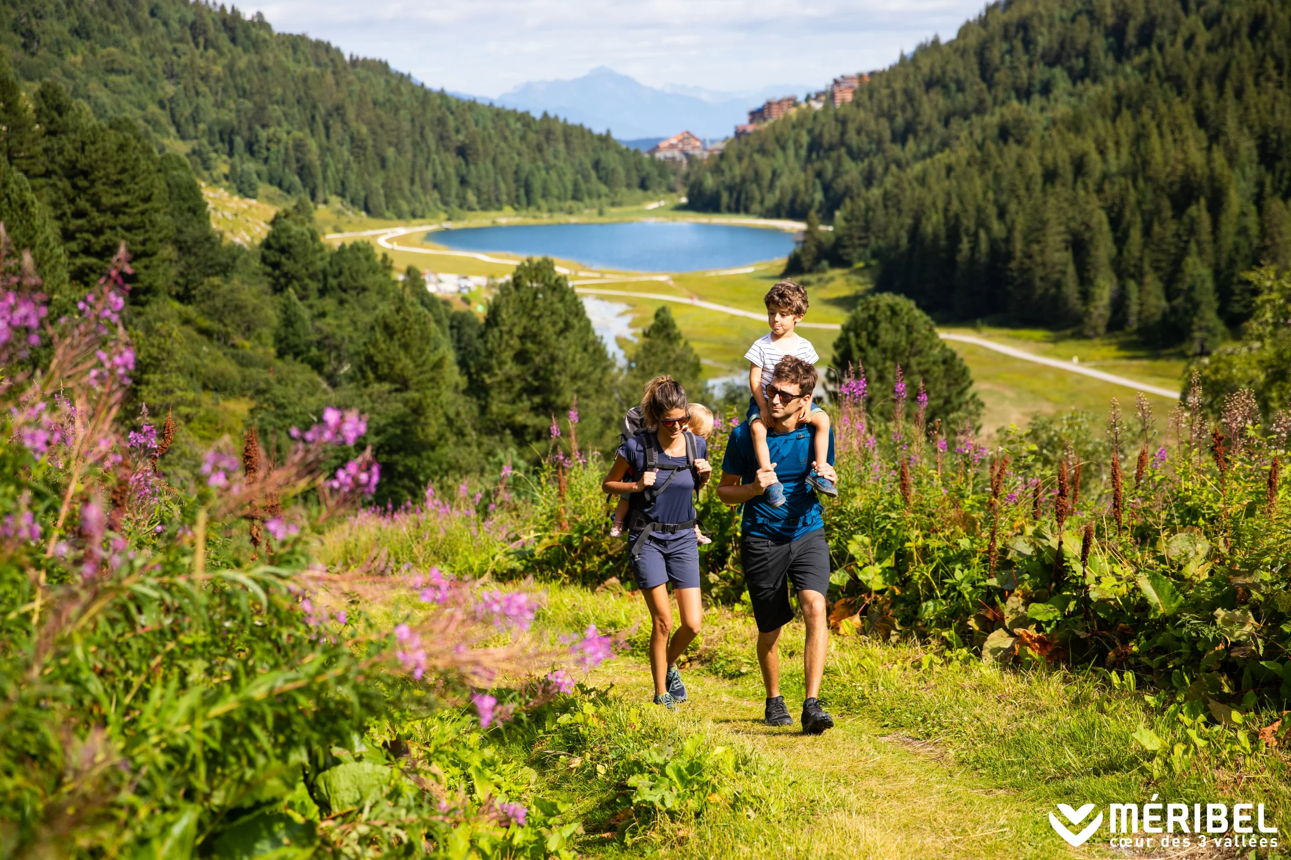 Randonnée en famille à Méribel sur un sentier fleuri au-dessus d’un lac, avec deux enfants portés par les adultes.