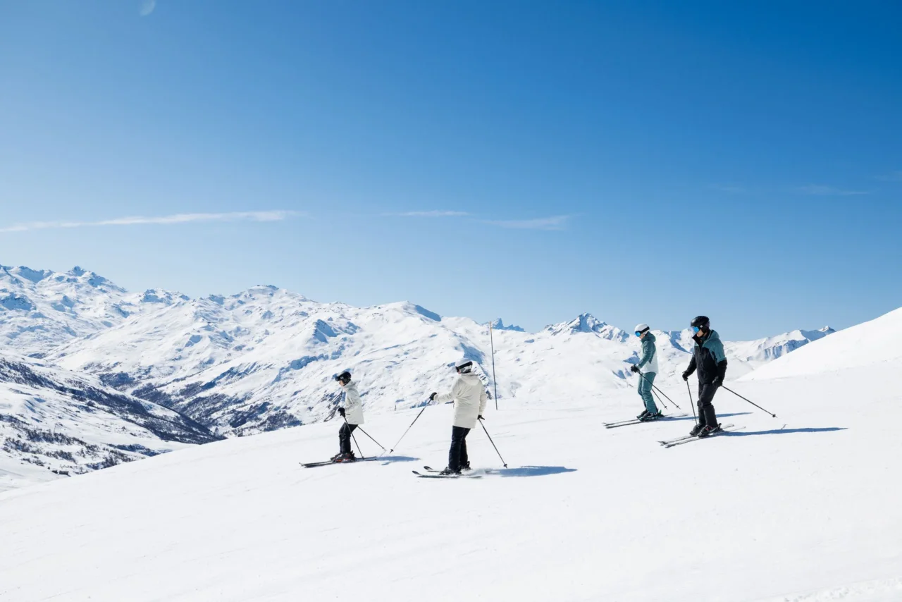 Quatre skieurs sur une piste enneigée face à un panorama de montagnes sous un ciel bleu.