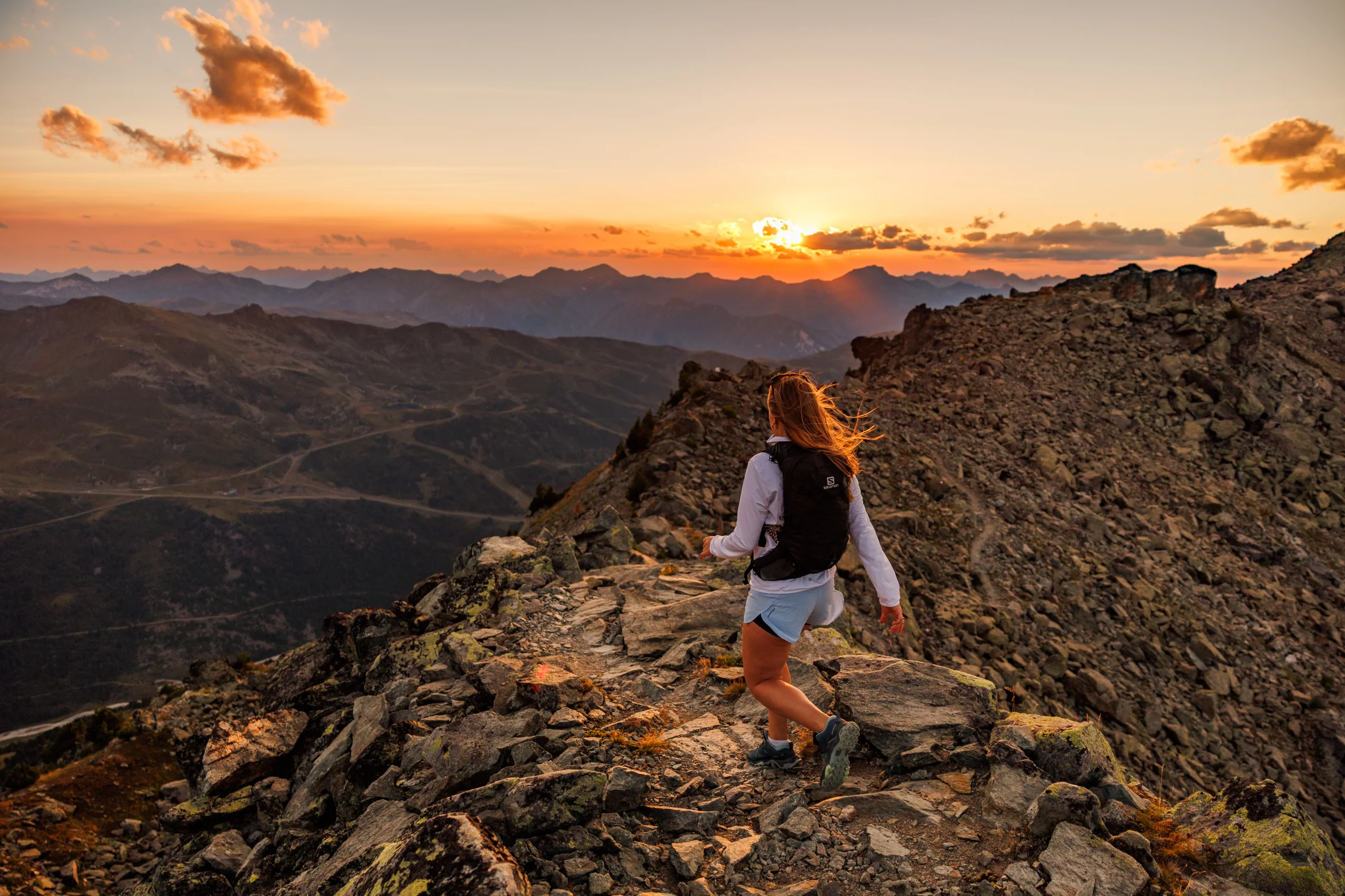 Une randonneuse avec un sac à dos avance sur un sentier rocheux au coucher du soleil.