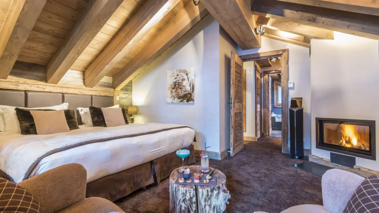 Chambre de l’Hôtel La Chaudanne sous une charpente apparente, avec un grand lit et une cheminée allumée.