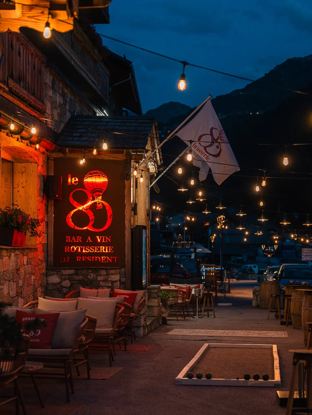Terrasse du bar à vin et rôtisserie Le 80 à la tombée de la nuit, éclairée de guirlandes, avec fauteuils et piste de pétanque.