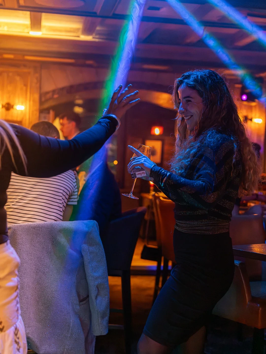 Une femme souriante danse, un verre à pied à la main, sous des faisceaux lumineux colorés.