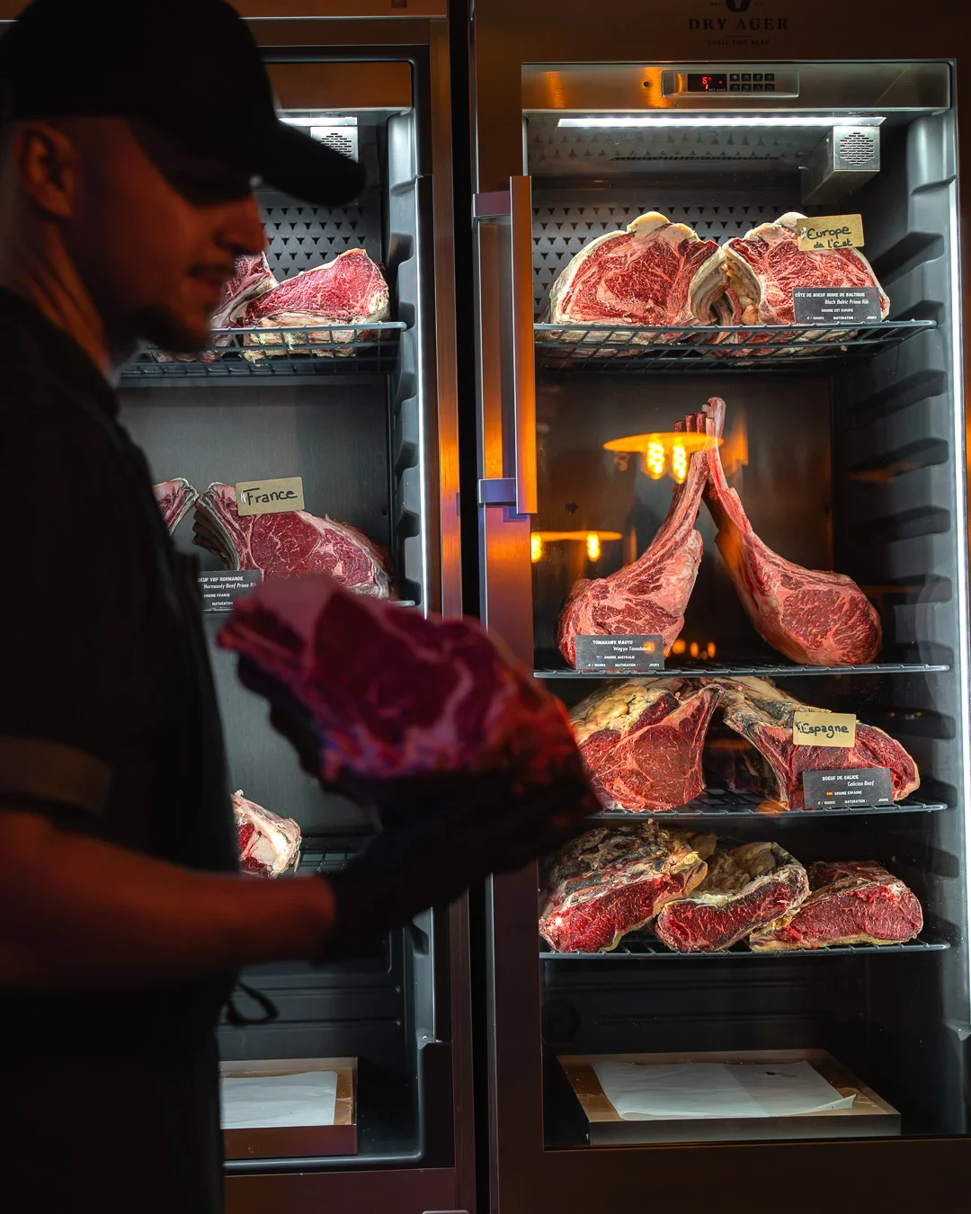 Une personne tient une pièce de bœuf devant des armoires de maturation garnies de côtes et de tomahawks.