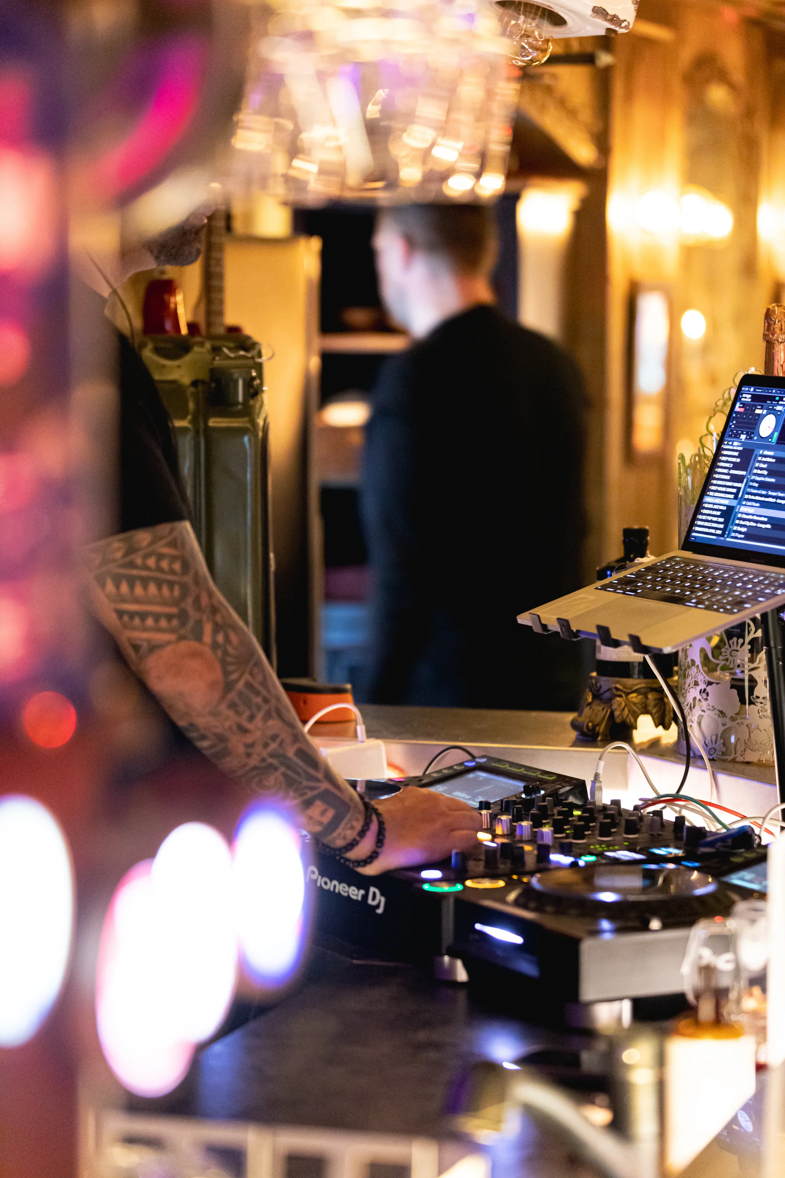 Un DJ au bras tatoué manipule une console de mixage reliée à un ordinateur portable.