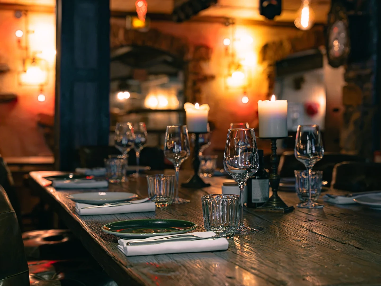Table de restaurant en bois dressée avec assiettes, verres à pied et bougies allumées.