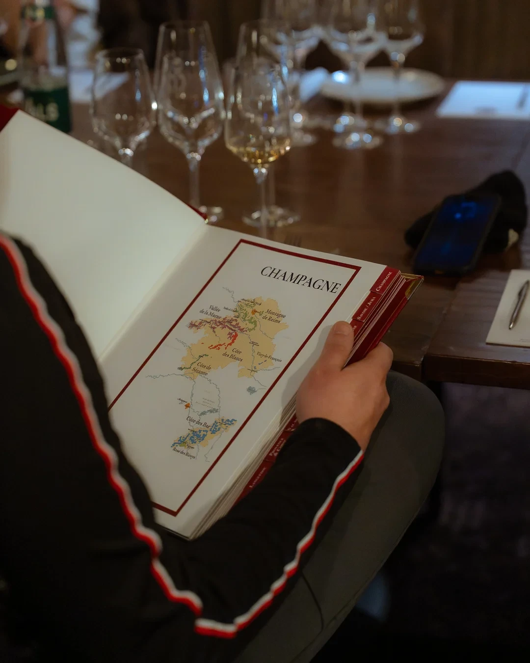 Une personne consulte un ouvrage ouvert sur une carte des vignobles de Champagne, devant des verres de dégustation.