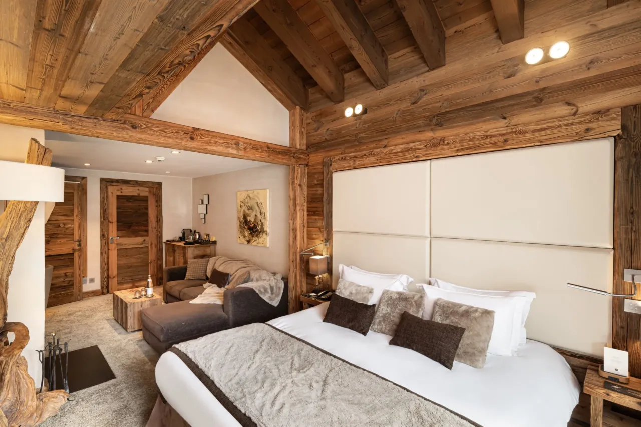 Chambre aux poutres apparentes avec lit double, grande tête de lit capitonnée et coin salon avec canapé.