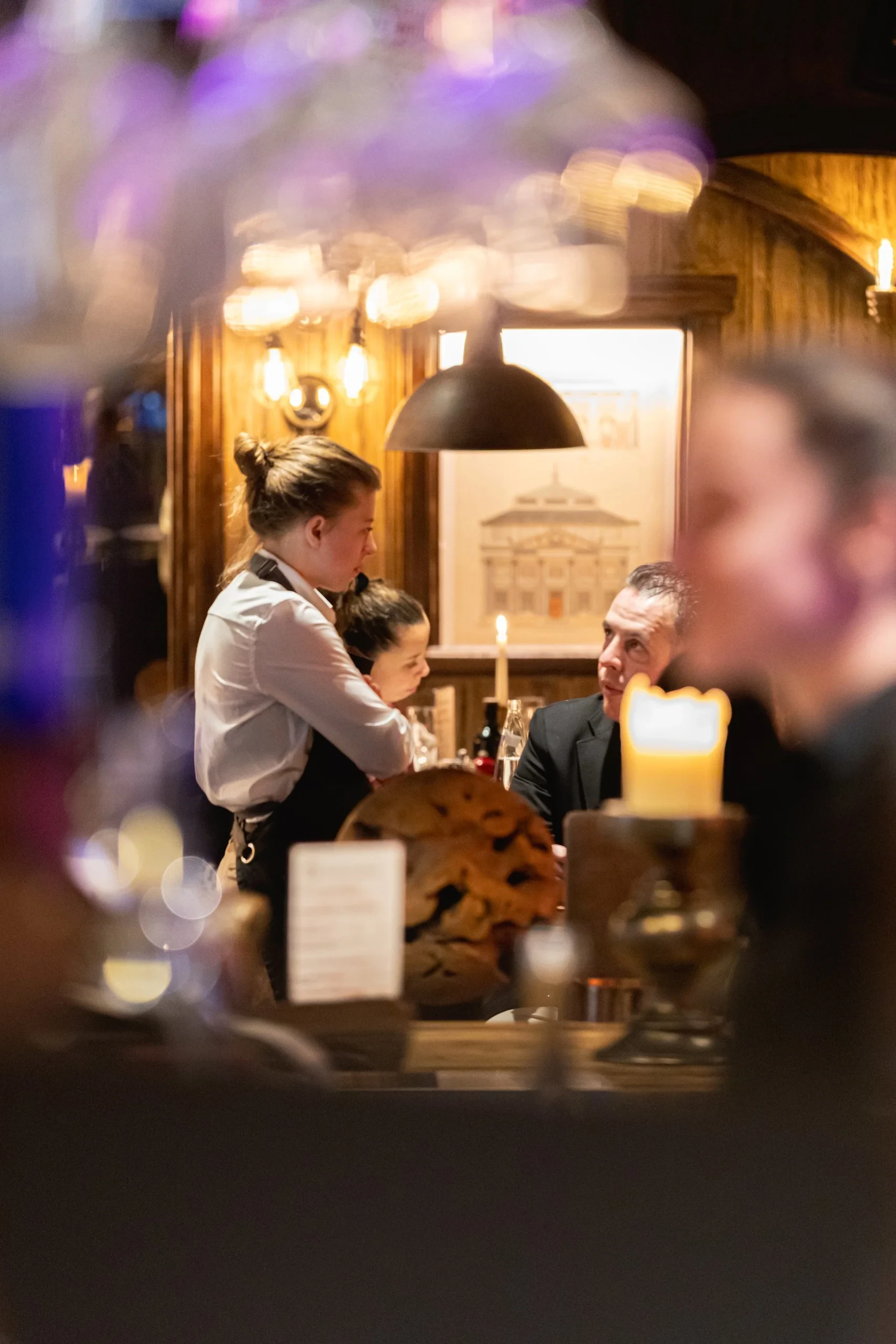 Une serveuse échange avec un client attablé dans une salle de restaurant aux boiseries éclairées par des lampes et des bougies.