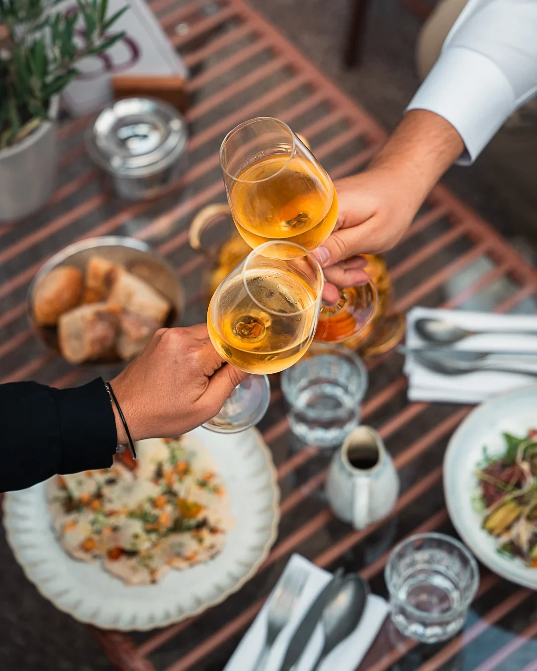 Deux personnes trinquent avec des verres de vin blanc au-dessus d’une table garnie de plats.