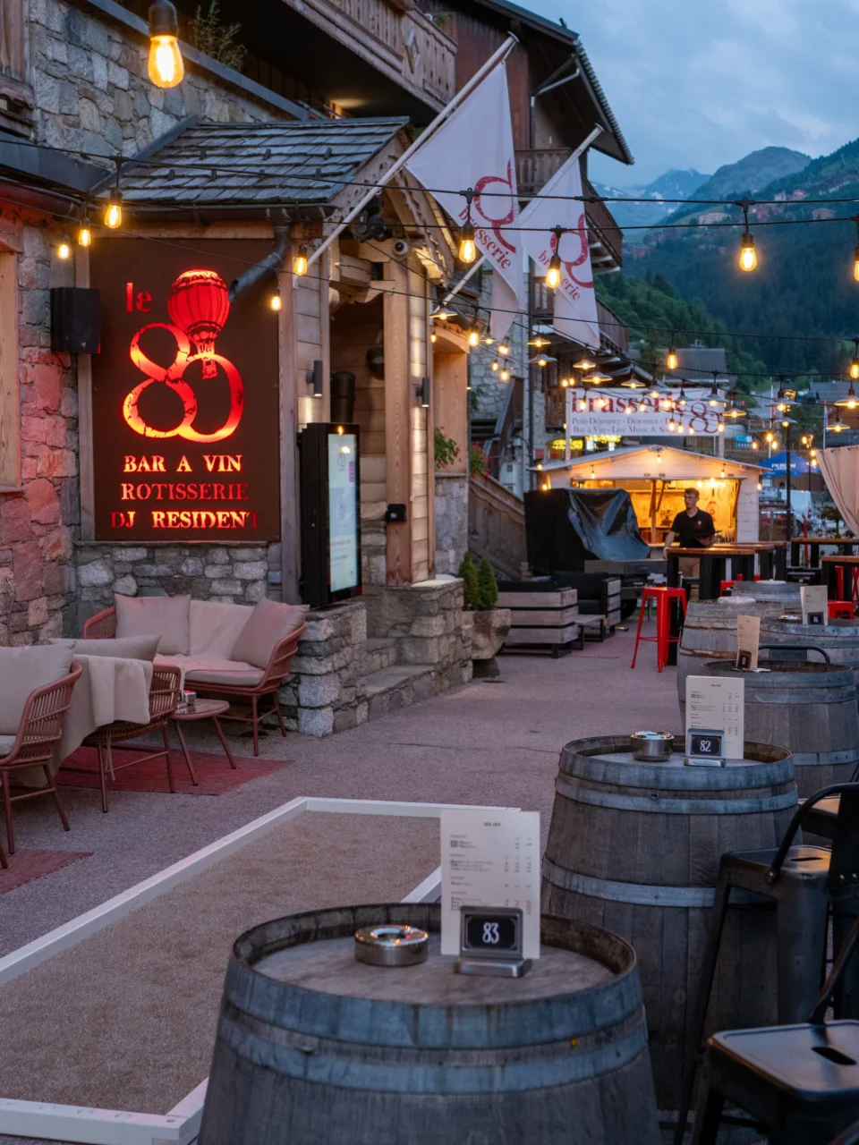 Terrasse du 80 au crépuscule, avec tables en tonneaux et guirlandes lumineuses devant les montagnes.