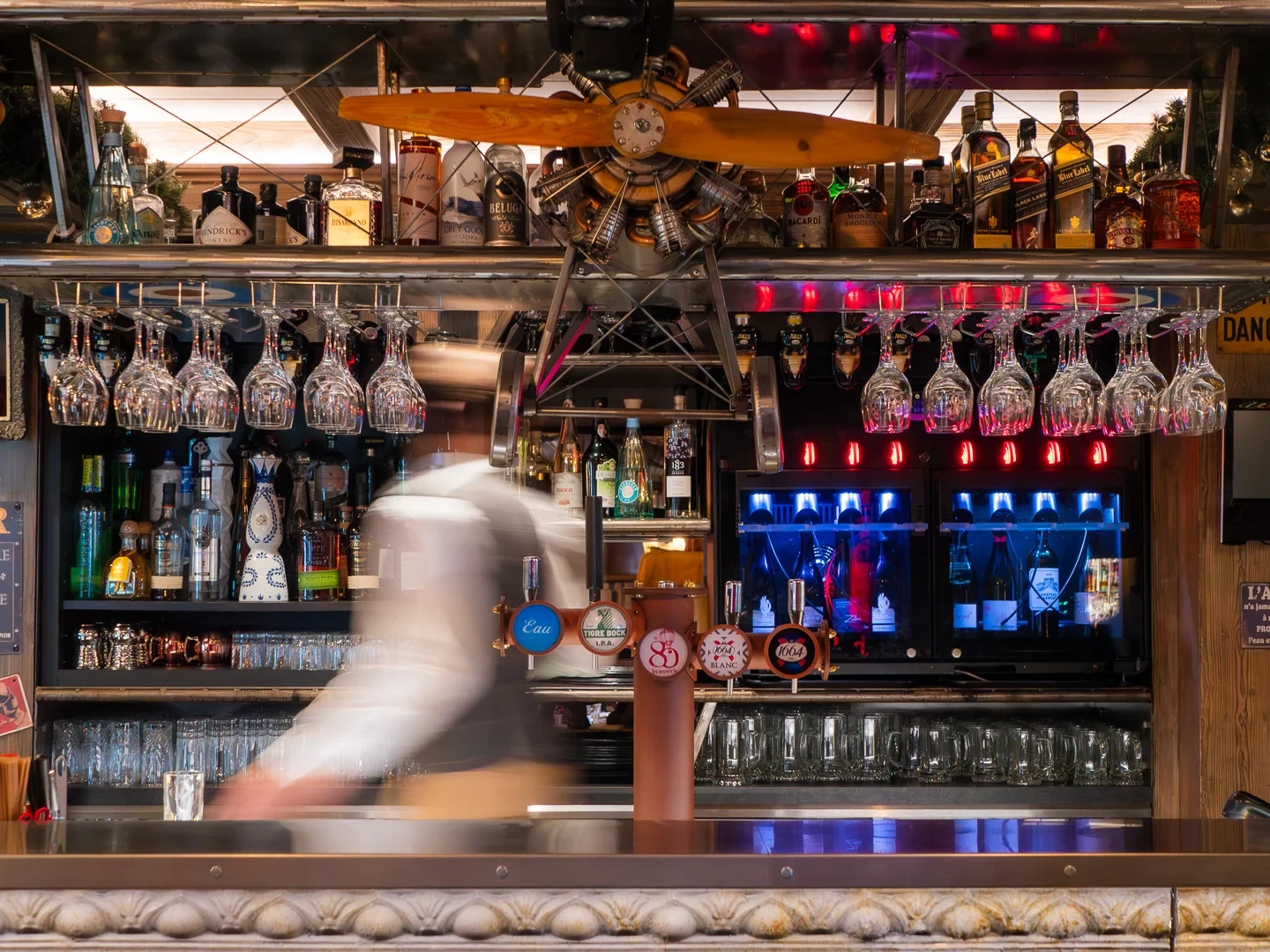 Comptoir de bar surmonté d’une hélice en bois, avec un barman flou en mouvement derrière les tireuses.