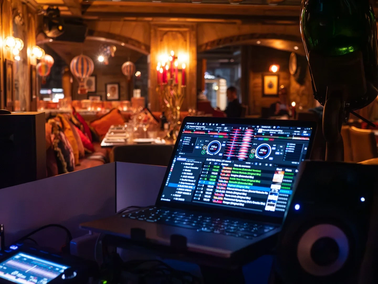 Installation de DJ avec ordinateur et enceinte devant une salle de restaurant aux boiseries et aux lumières tamisées.