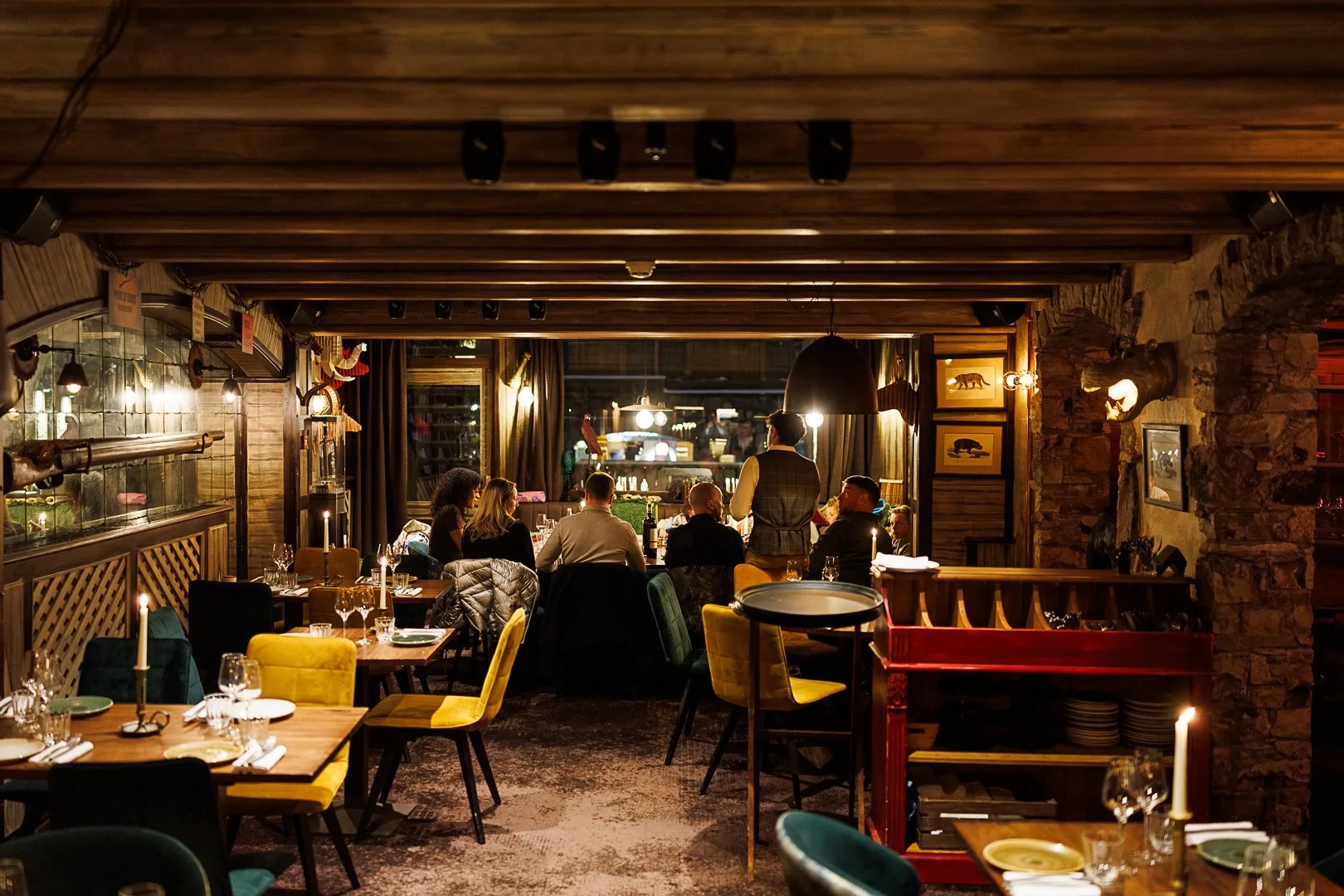 Salle de restaurant aux poutres apparentes, avec des tables éclairées à la bougie et des convives au fond.