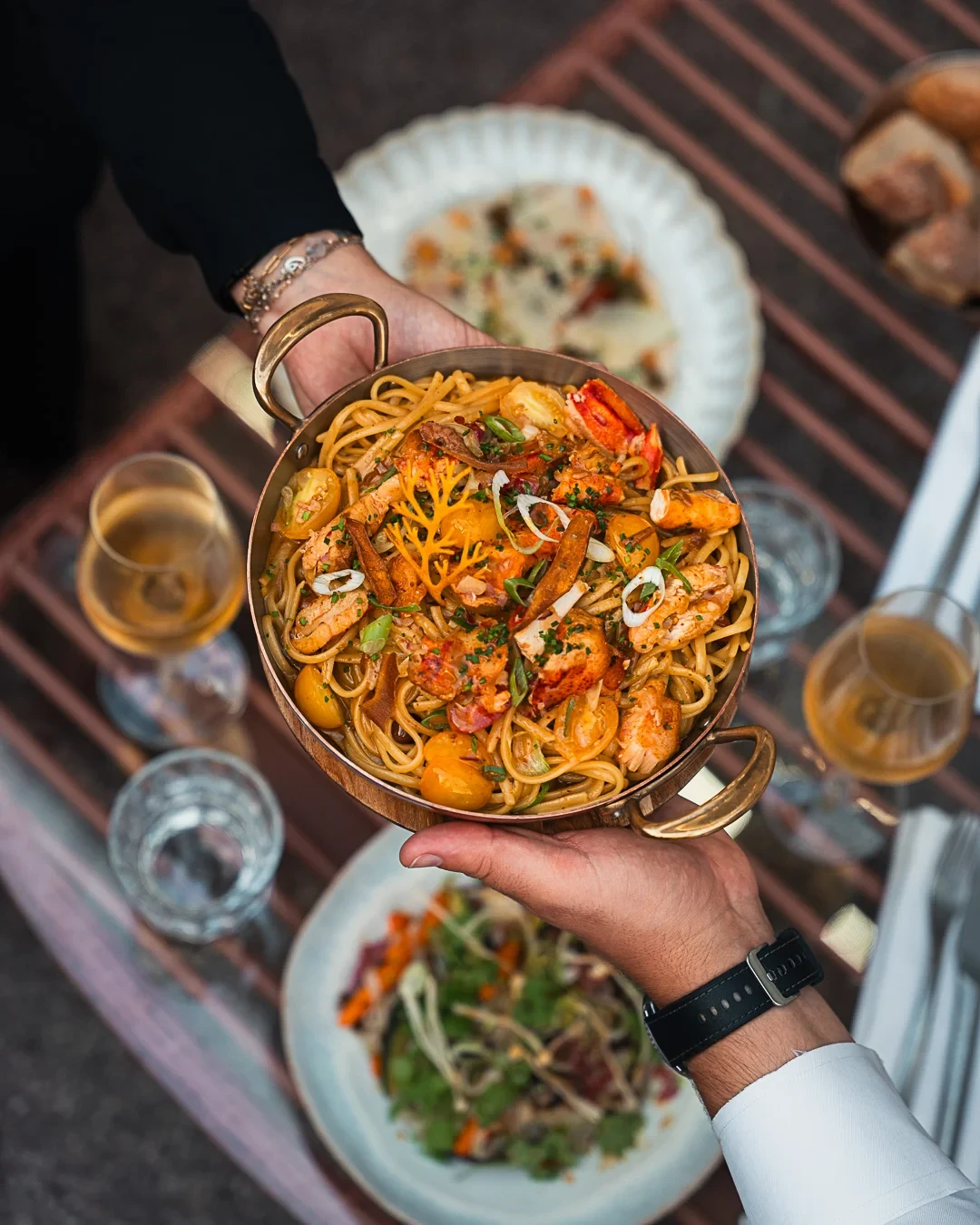 Plat de linguine au homard, garni de tomates jaunes et d’herbes, présenté à deux mains.