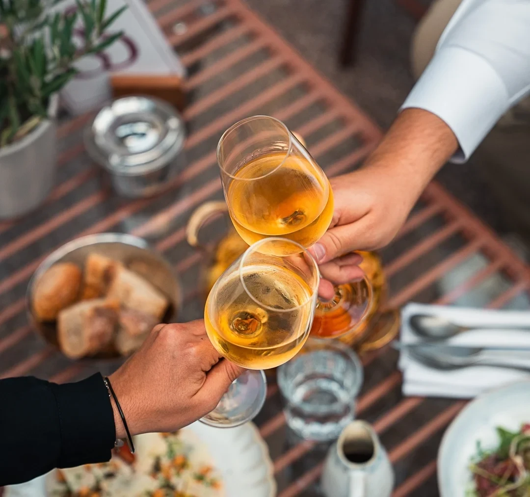Deux personnes trinquent avec des verres de vin blanc au-dessus d’une table dressée.