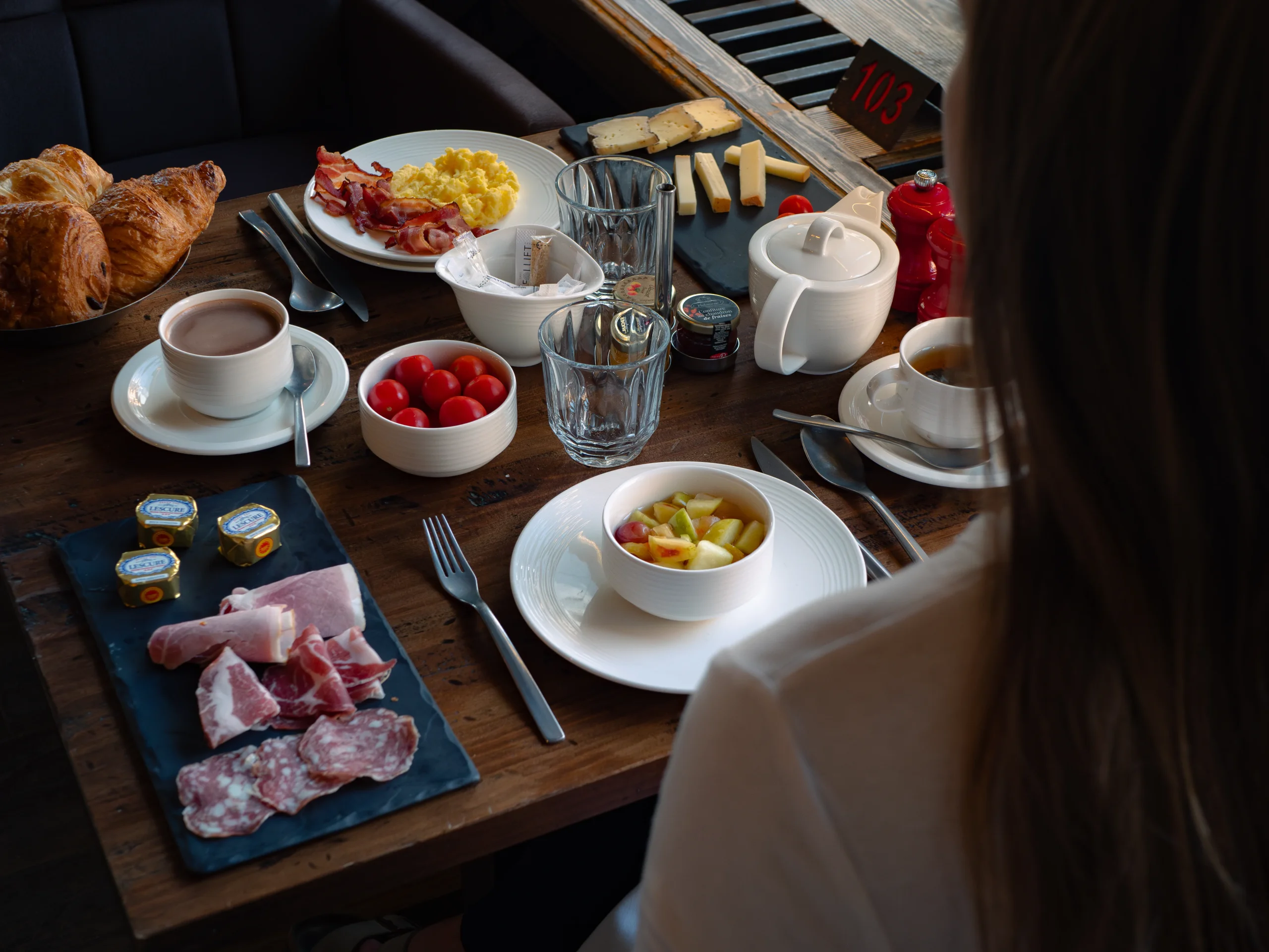 Petit-déjeuner servi à table avec viennoiseries, œufs brouillés, bacon, fromages, charcuterie et salade de fruits.