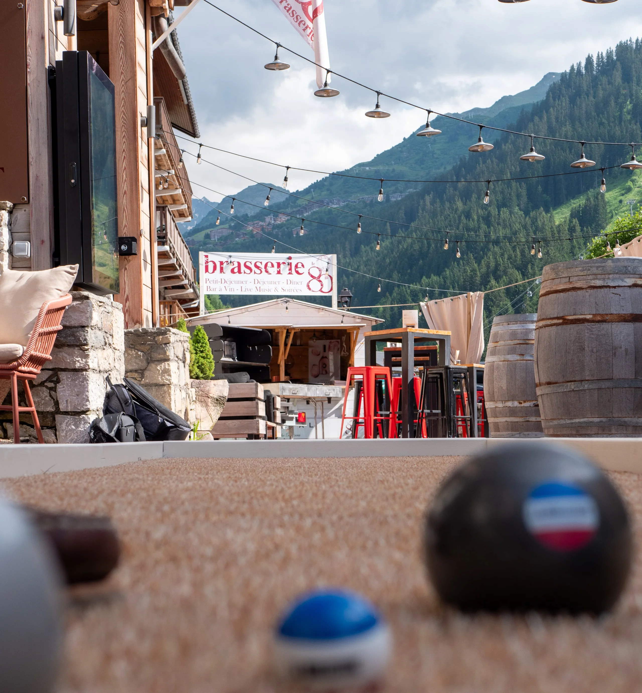 Terrain de pétanque devant la terrasse de la brasserie Le 80, avec les montagnes boisées en arrière-plan.