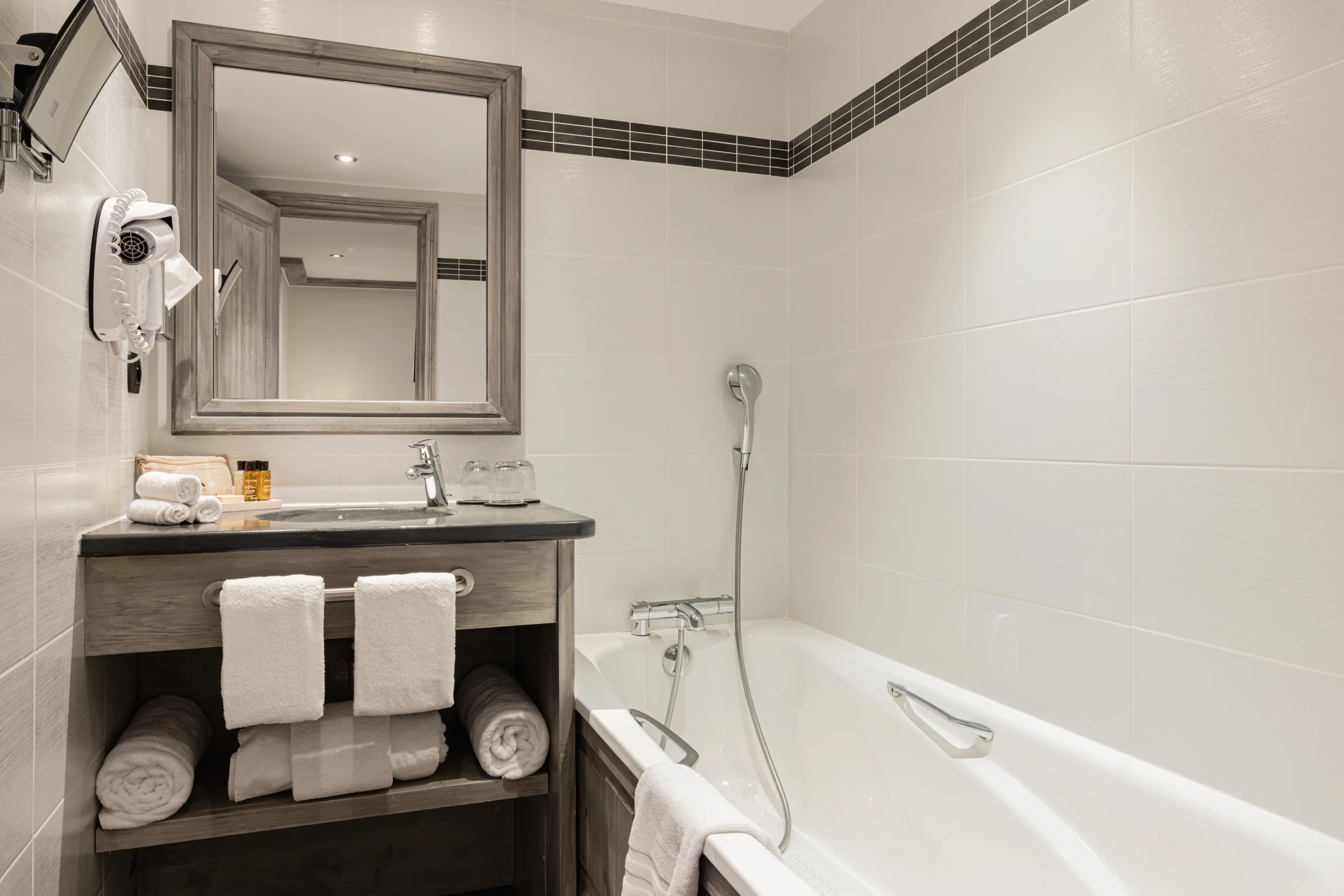 Salle de bains de l’Hôtel La Chaudanne avec baignoire, douchette et vasque sur un meuble en bois grisé.