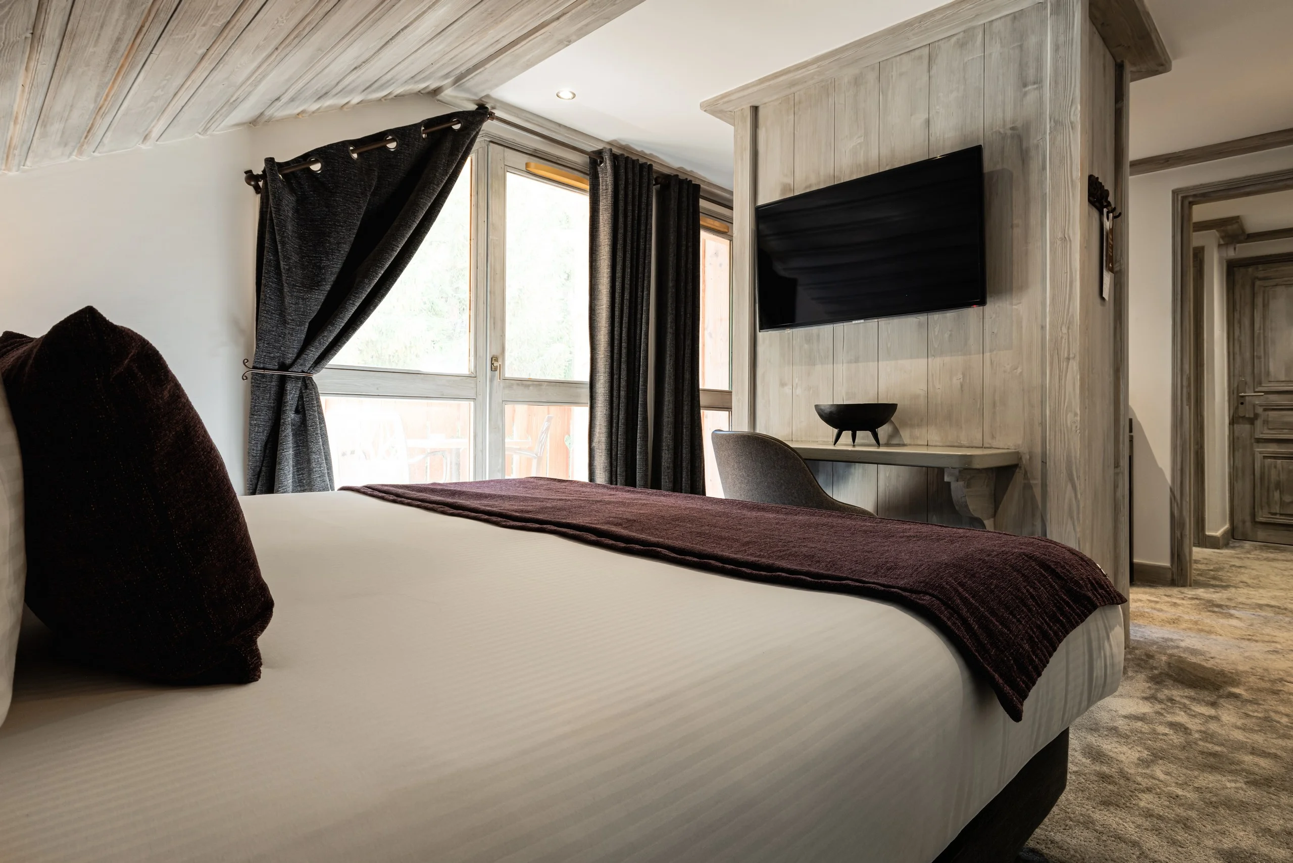 Chambre aux boiseries claires avec lit double, grandes fenêtres à rideaux sombres et télévision murale.
