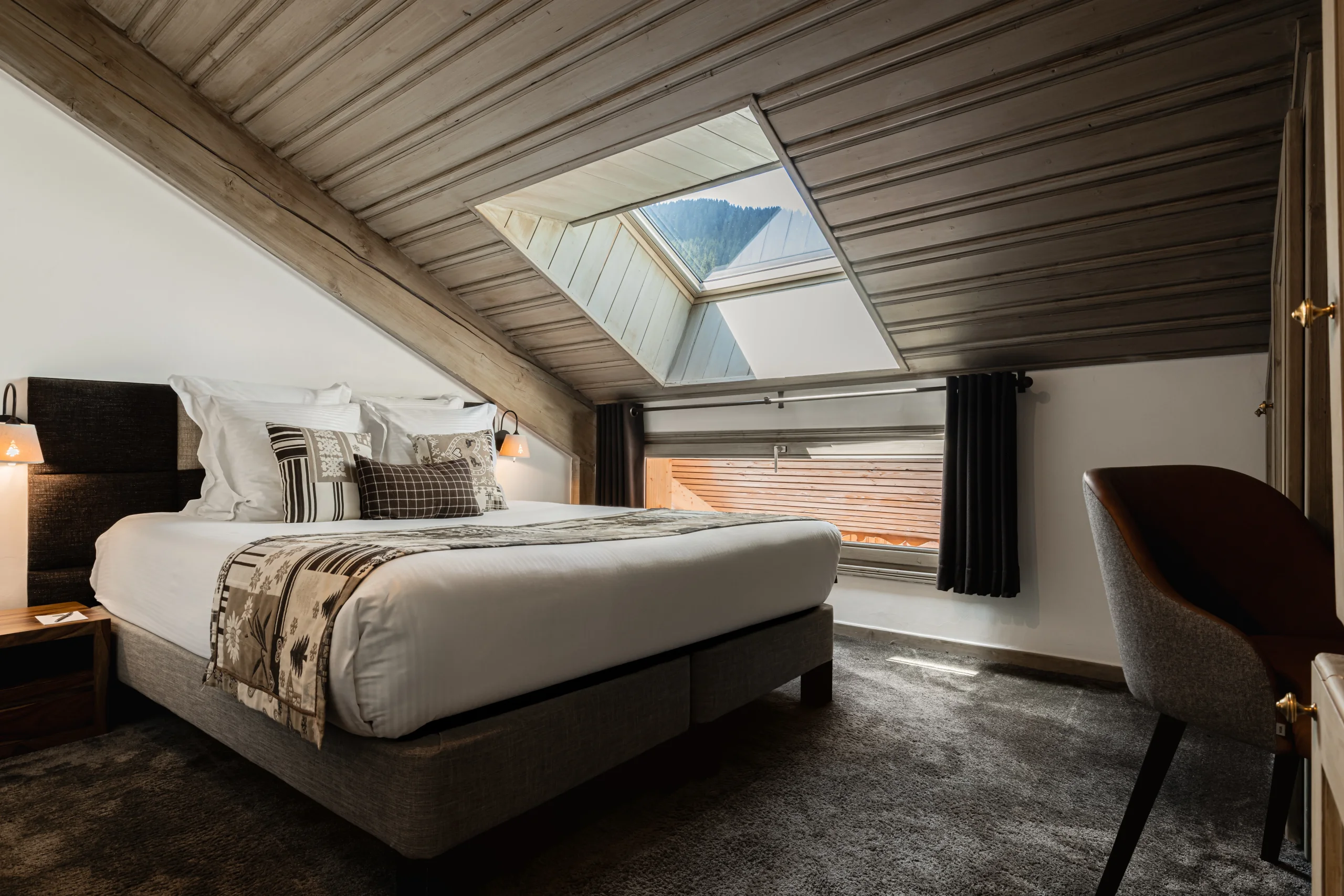 Chambre mansardée avec lit double sous un plafond en bois et fenêtre de toit ouvrant sur la montagne.