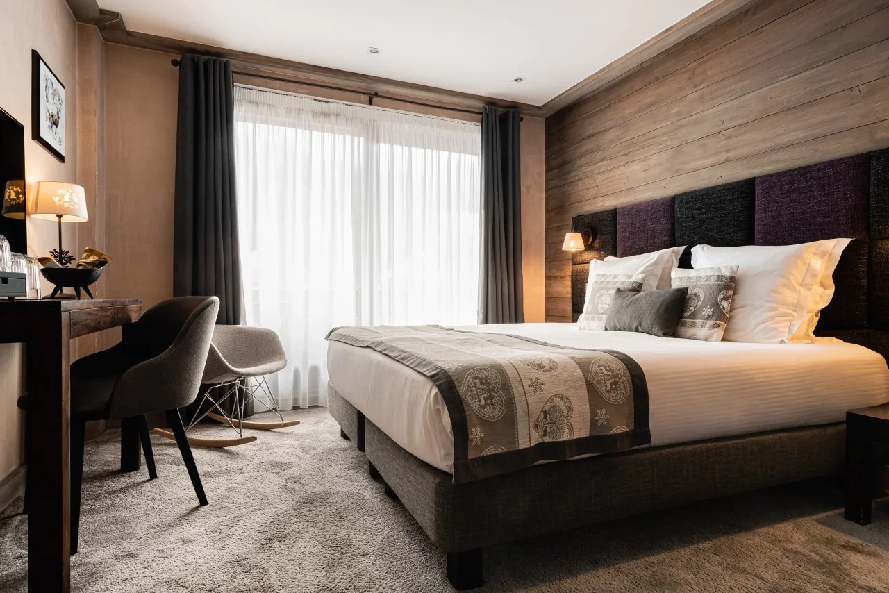 Chambre de l’Hôtel La Chaudanne avec lit double, tête de lit capitonnée et fauteuil à bascule près de la baie vitrée.
