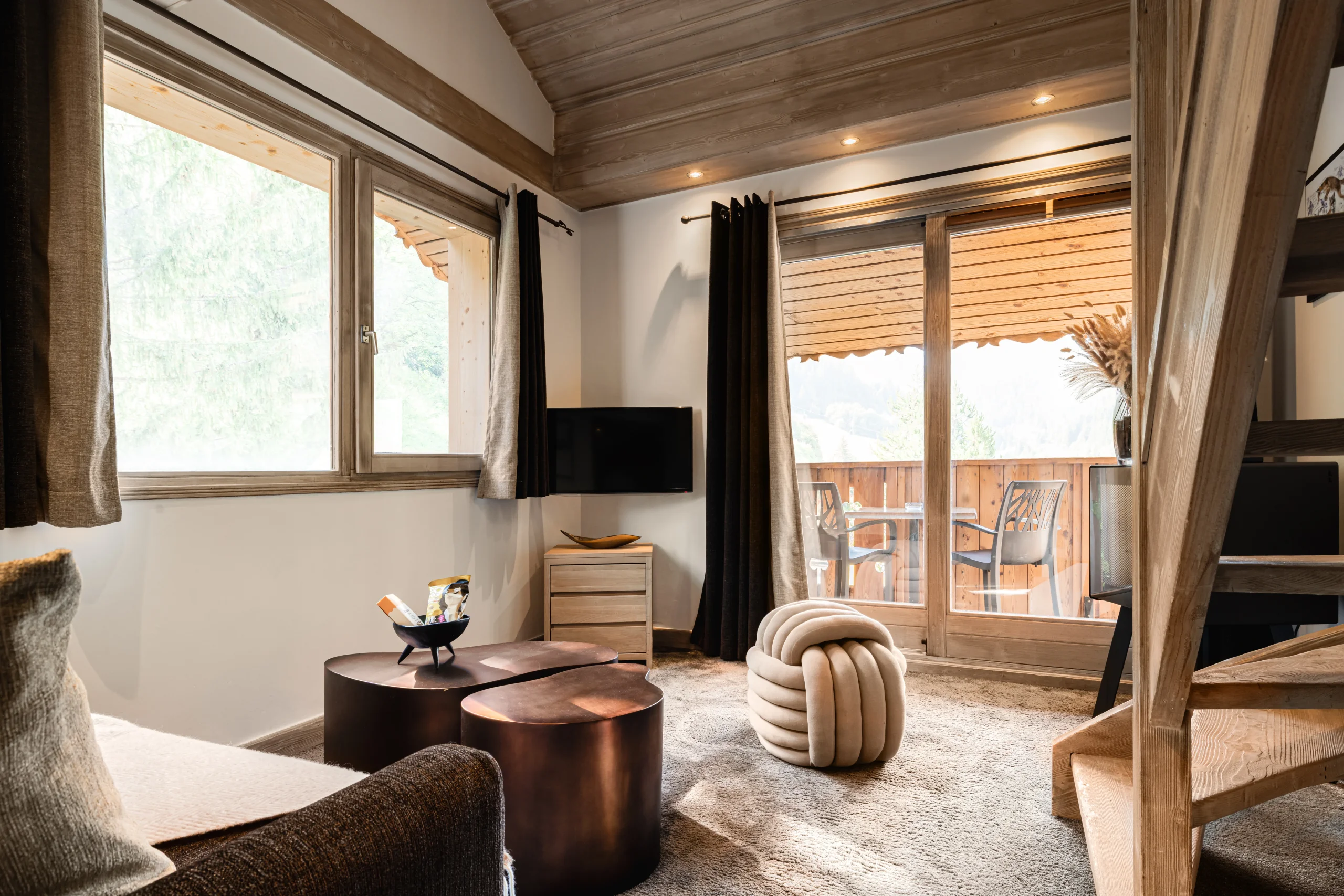 Salon avec accès à un balcon aménagé, tables basses et pouf noué près d’un escalier en bois.
