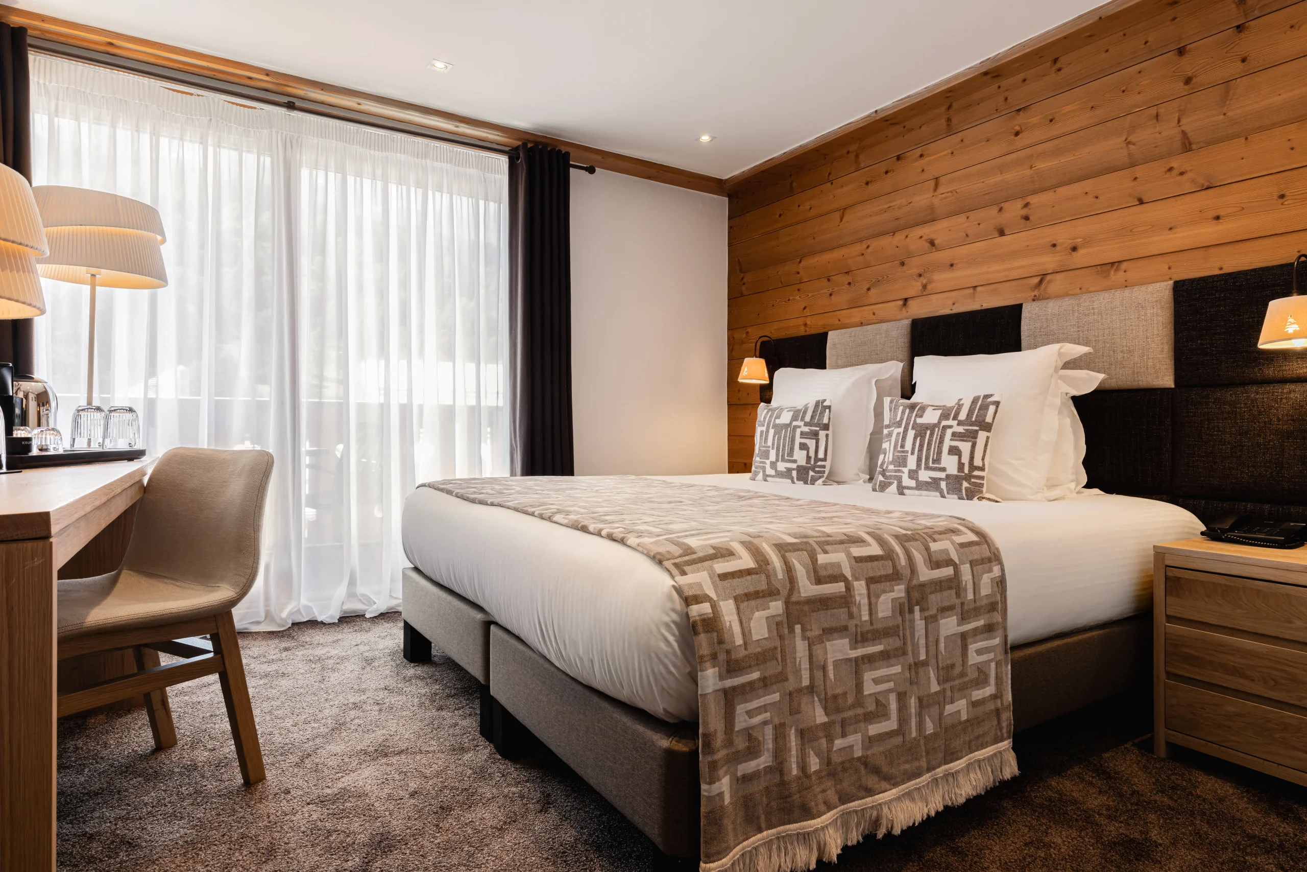 Chambre de l’Hôtel La Chaudanne avec lit double, mur en bois et grande fenêtre voilée.
