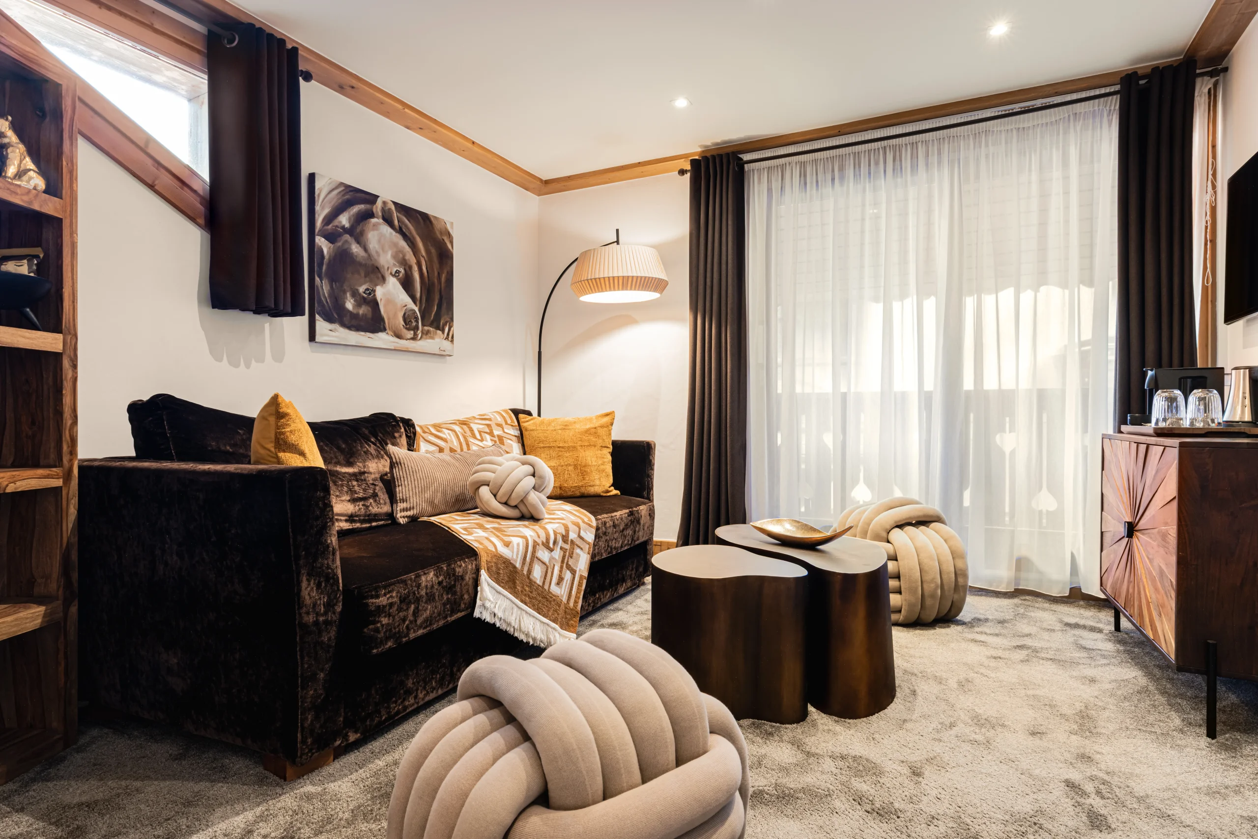 Salon de l’Hôtel La Chaudanne avec canapé brun, coussins ocre et poufs en forme de nœuds devant une grande baie vitrée.