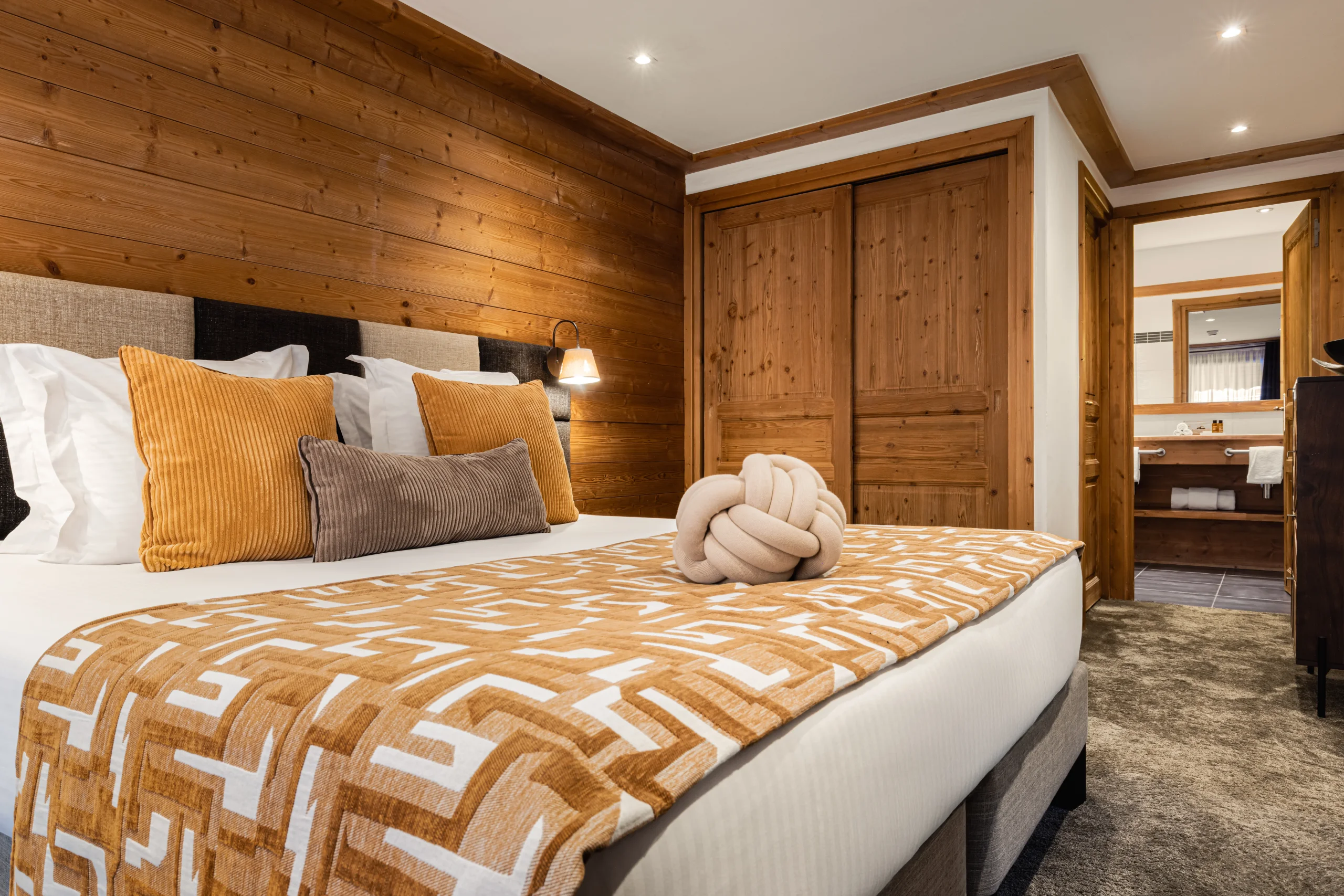 Lit double avec plaid à motifs ocre, face aux portes de placard en bois et à la salle de bains ouverte.
