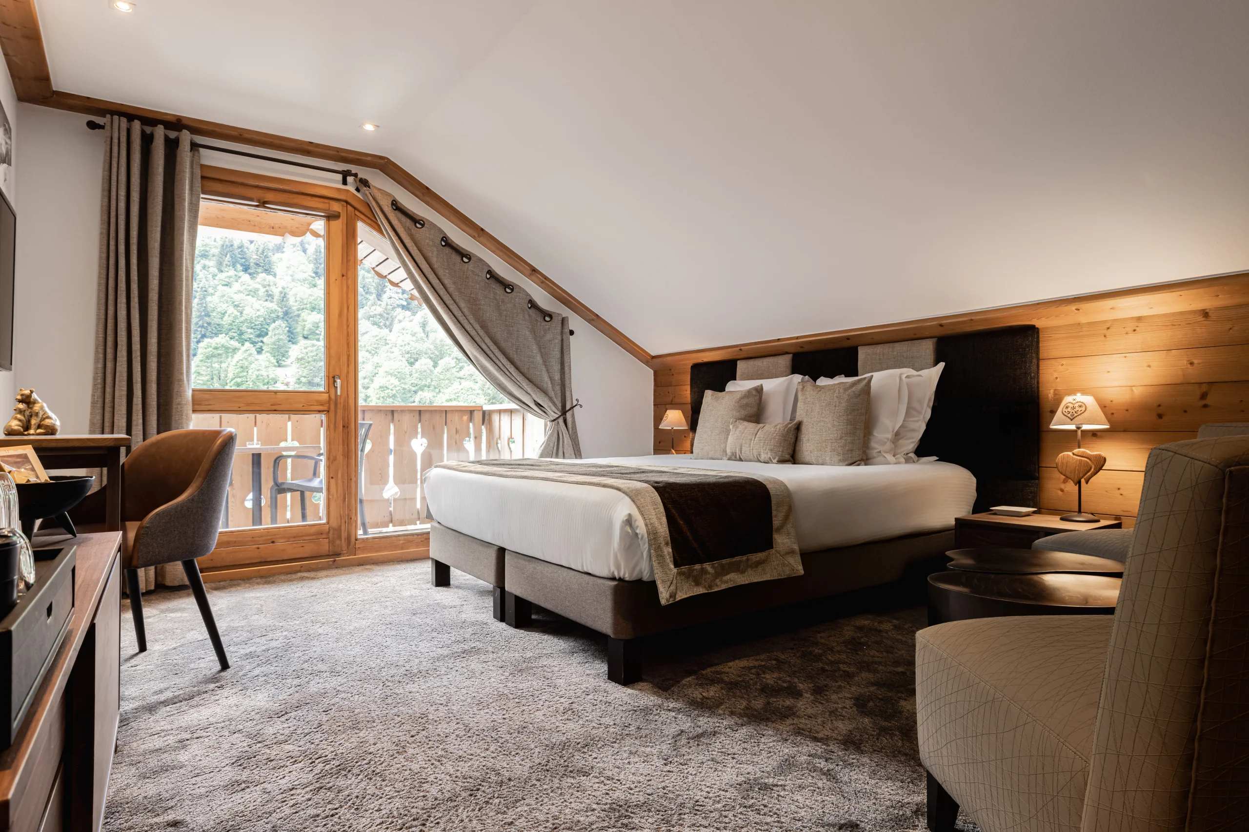 Chambre mansardée avec lit double et porte vitrée donnant sur un balcon face à un versant boisé.