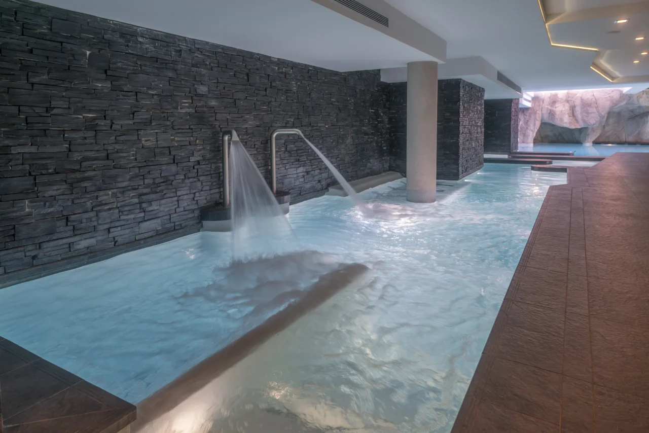 Deux jets d’eau en fonctionnement dans la piscine intérieure de l’Hôtel La Chaudanne, bordée de murs en pierre sombre.