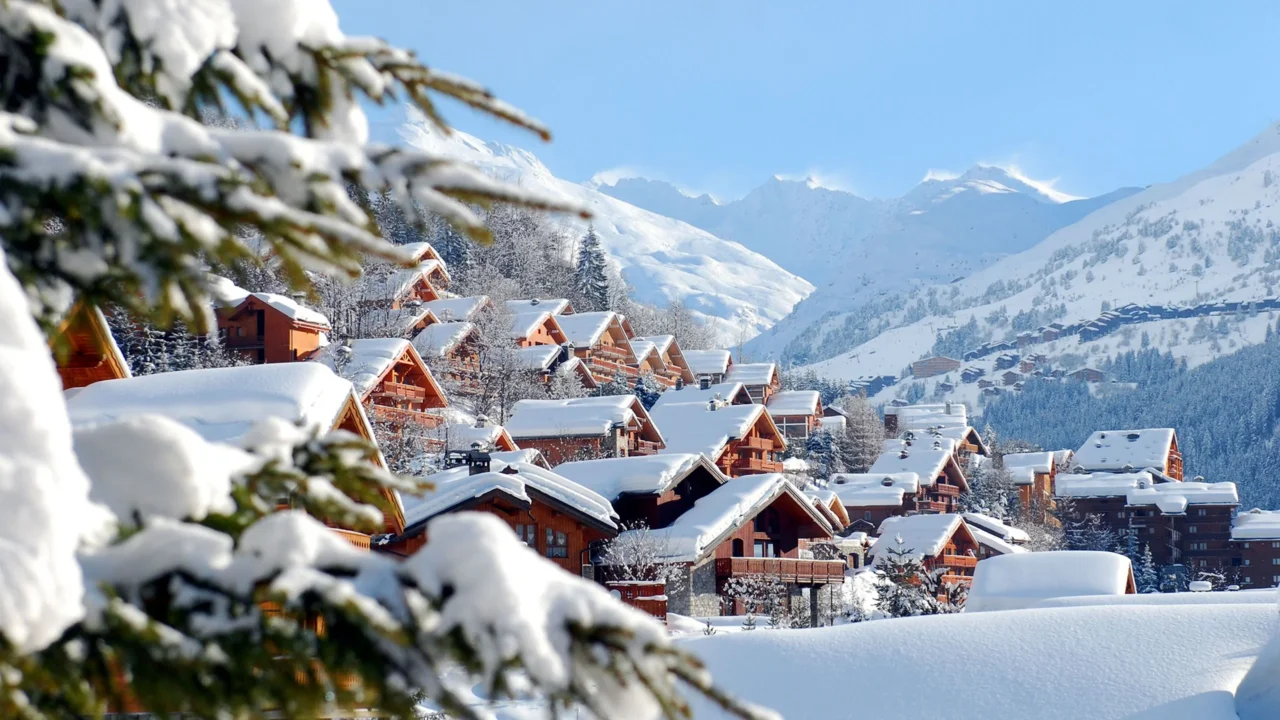 Chalets en bois couverts de neige sur un versant, face aux sommets enneigés.