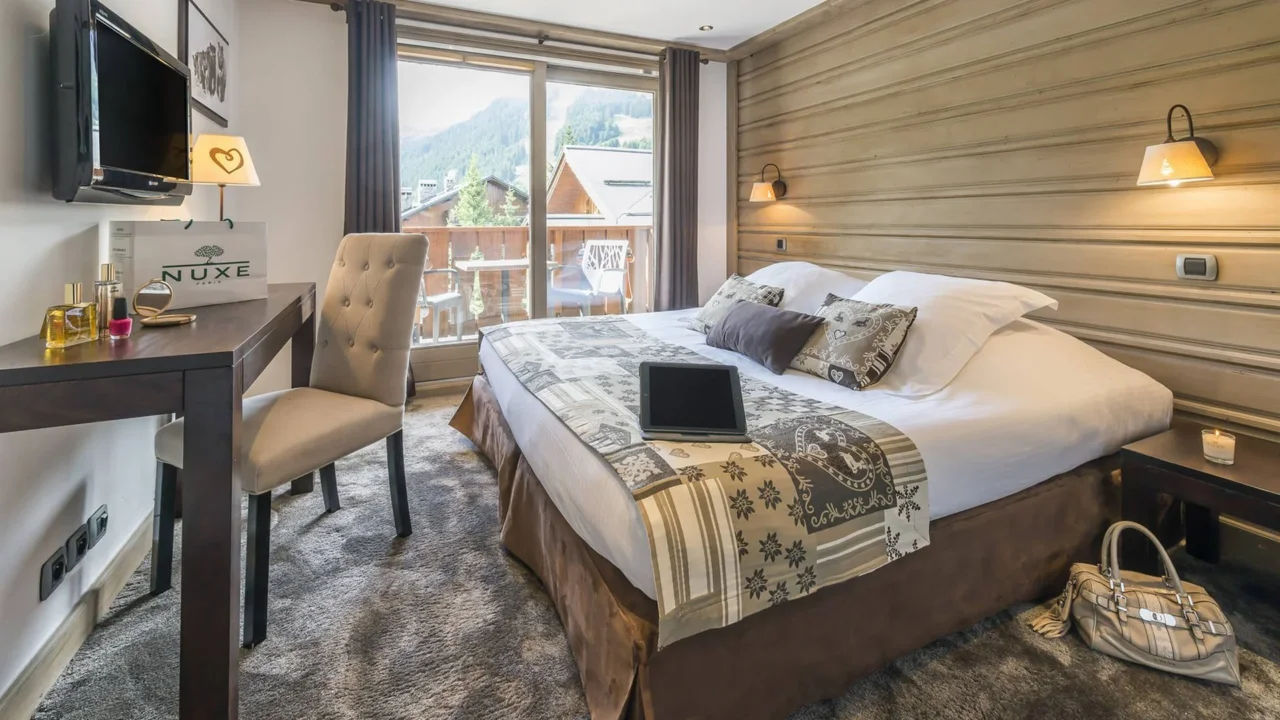 Chambre avec lit double, bureau et balcon donnant sur des chalets et les montagnes.