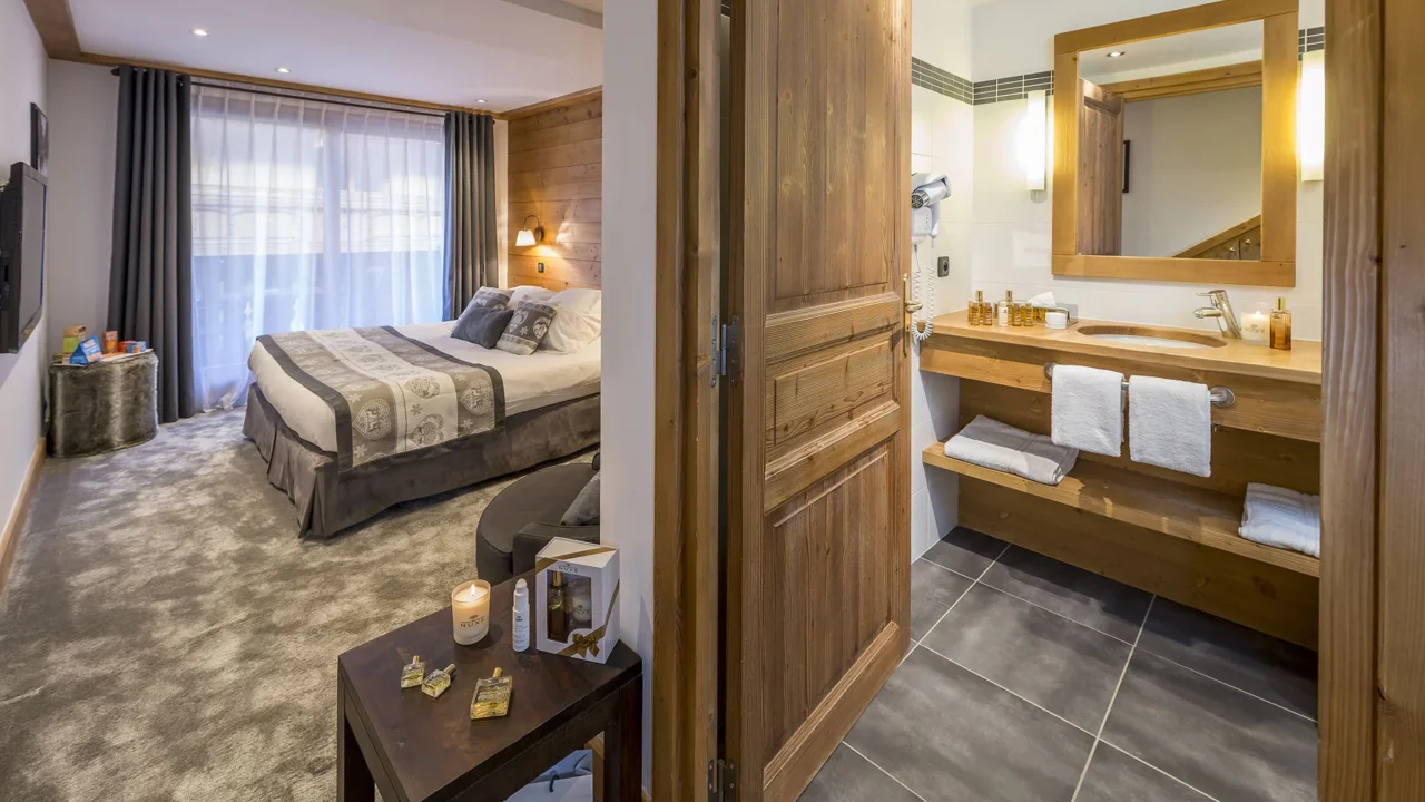 Chambre avec lit double et parement mural en bois, à côté d’une salle de bain dont la porte est ouverte.