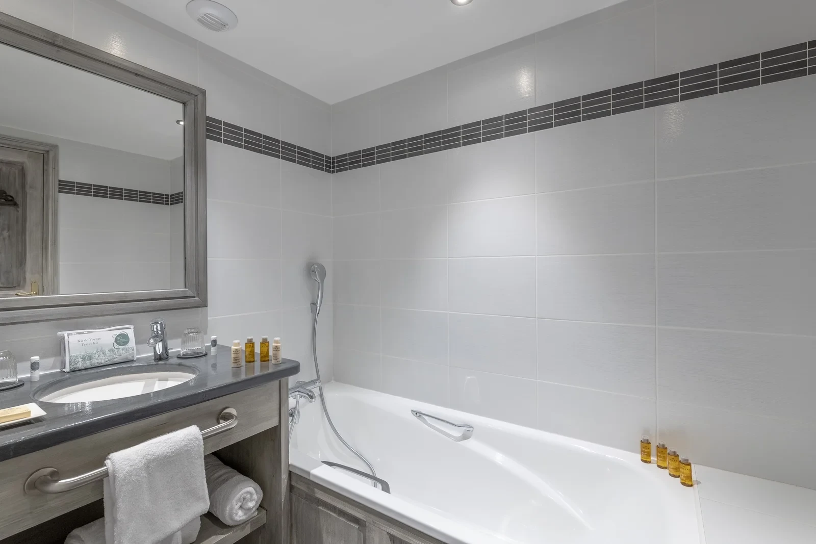 Salle de bain avec baignoire, douchette et vasque sur un meuble en bois grisé.
