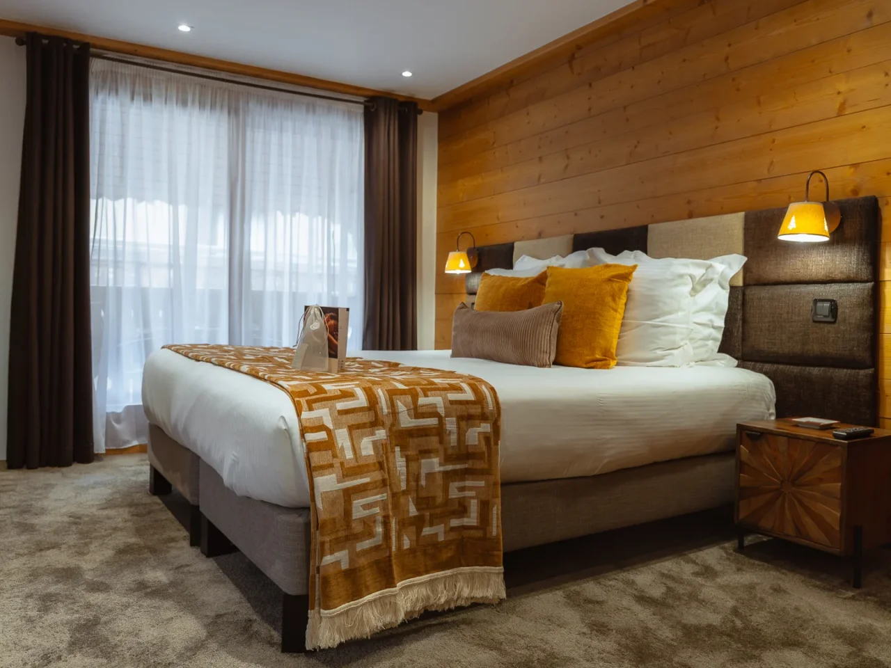 Chambre de l’Hôtel La Chaudanne avec lit double, mur en bois et coussins ocre.