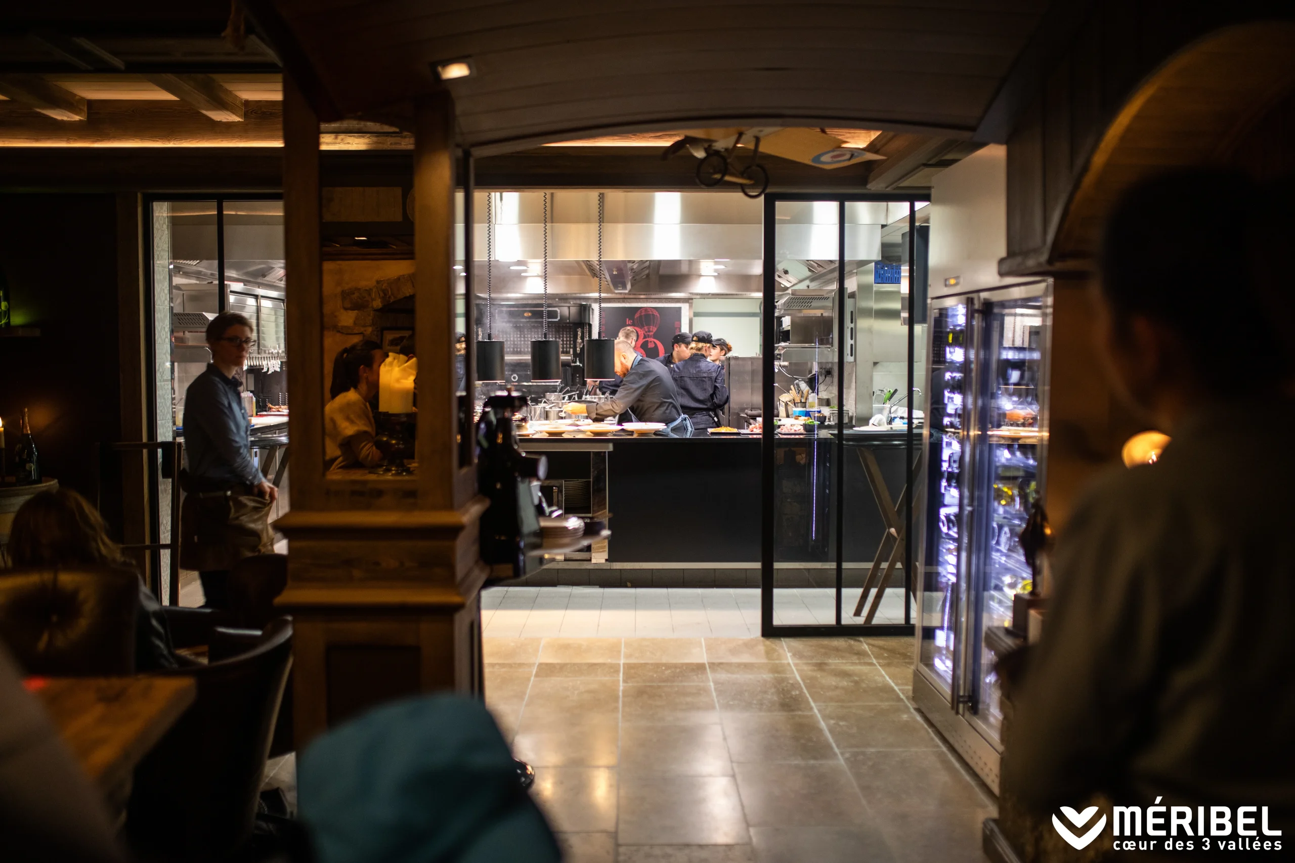 Une équipe travaille dans une cuisine ouverte sur une salle de restaurant aux boiseries sombres.