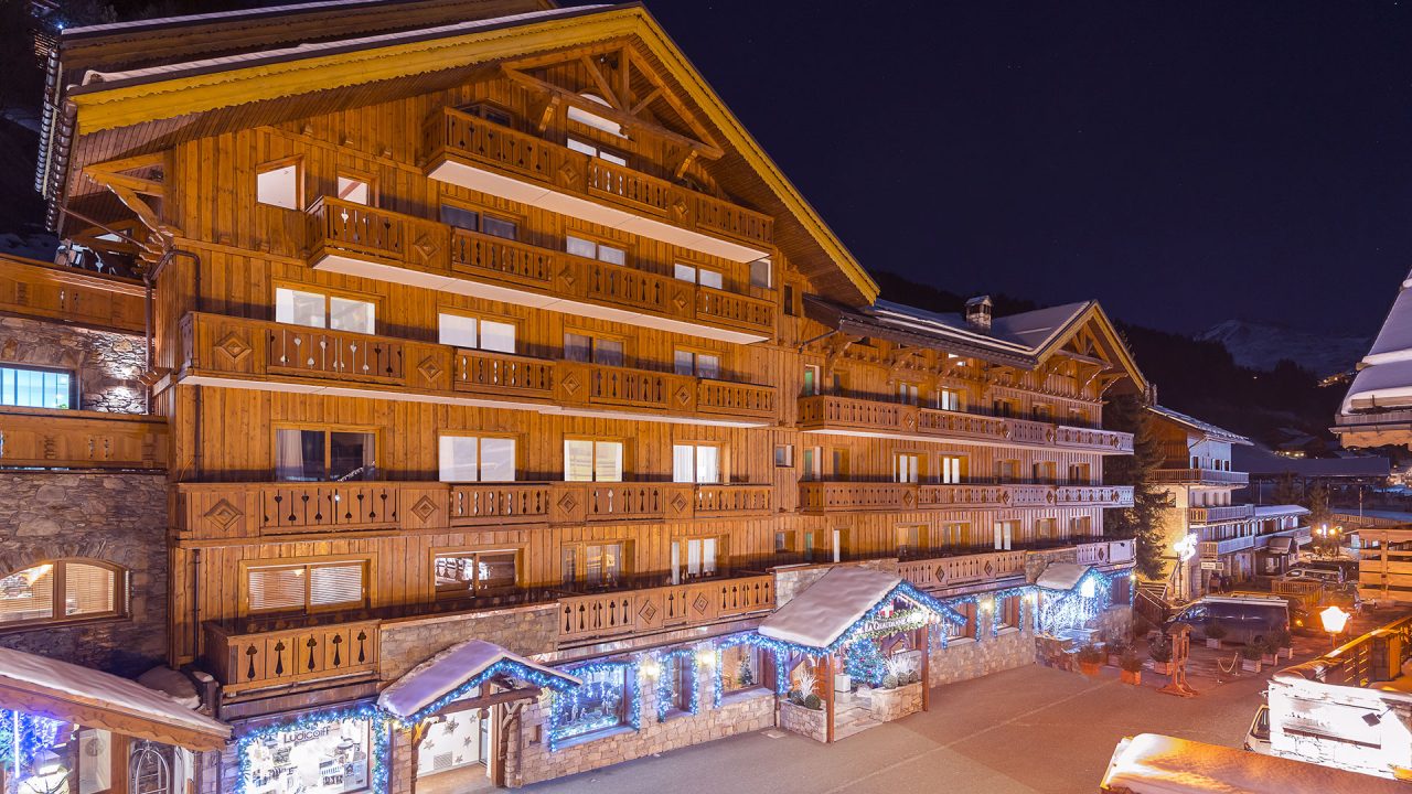 Façade en bois et balcons de l’Hôtel La Chaudanne illuminés de nuit, avec des décorations lumineuses sous les auvents enneigés.