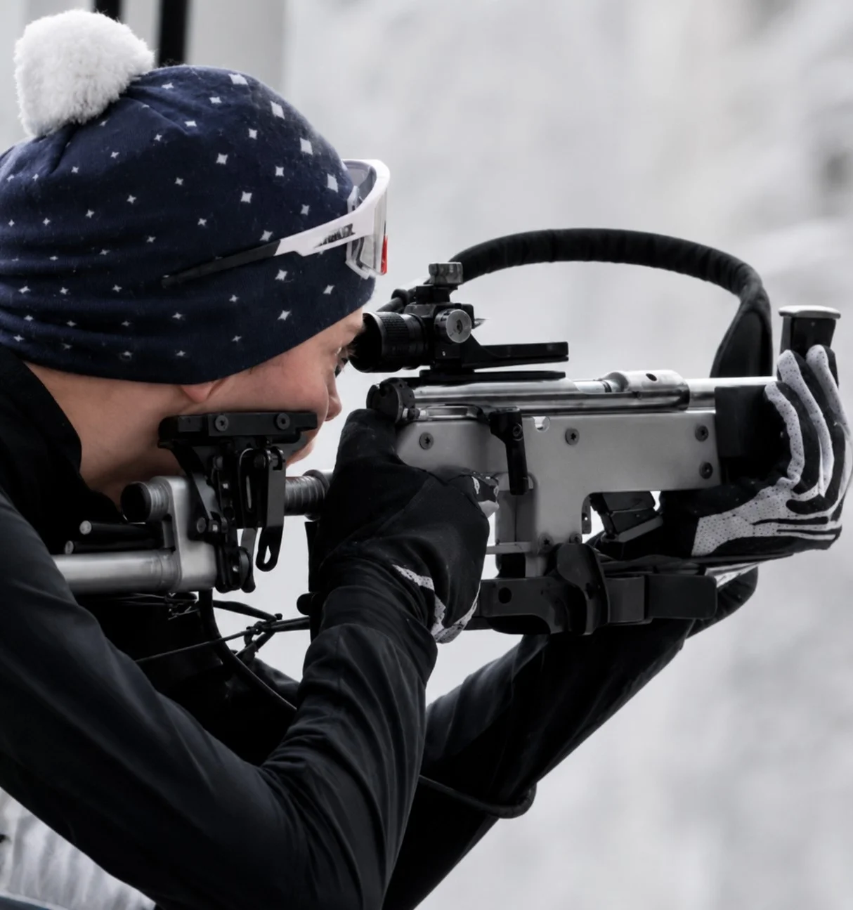 Personne en tenue de biathlon visant avec une carabine épaulée, sur fond enneigé.