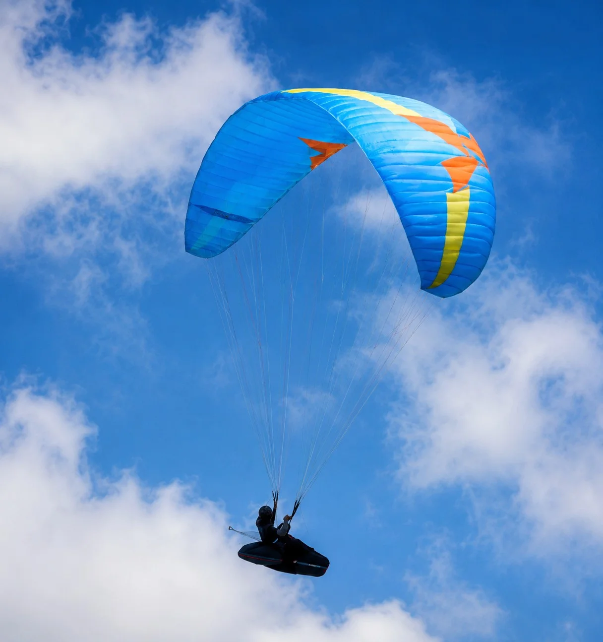 Parapentiste en vol sous une voile bleue, jaune et orange dans un ciel nuageux.
