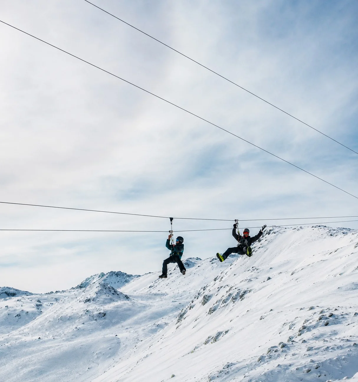 Deux personnes en tyrolienne sur des câbles parallèles au-dessus des montagnes enneigées.
