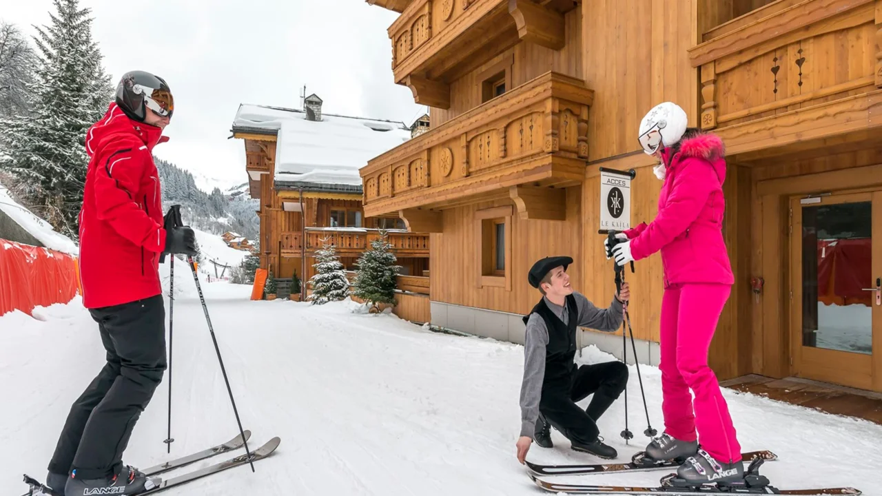 Un homme aide une skieuse à ajuster son équipement devant un chalet en bois, tandis qu’un autre skieur attend à proximité.