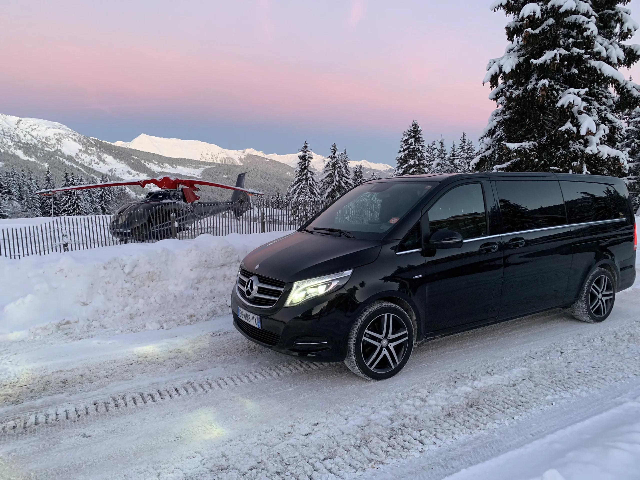 Van Mercedes noir vu de trois quarts avant sur une route enneigée, avec un hélicoptère derrière une clôture et les montagnes en arrière-plan.