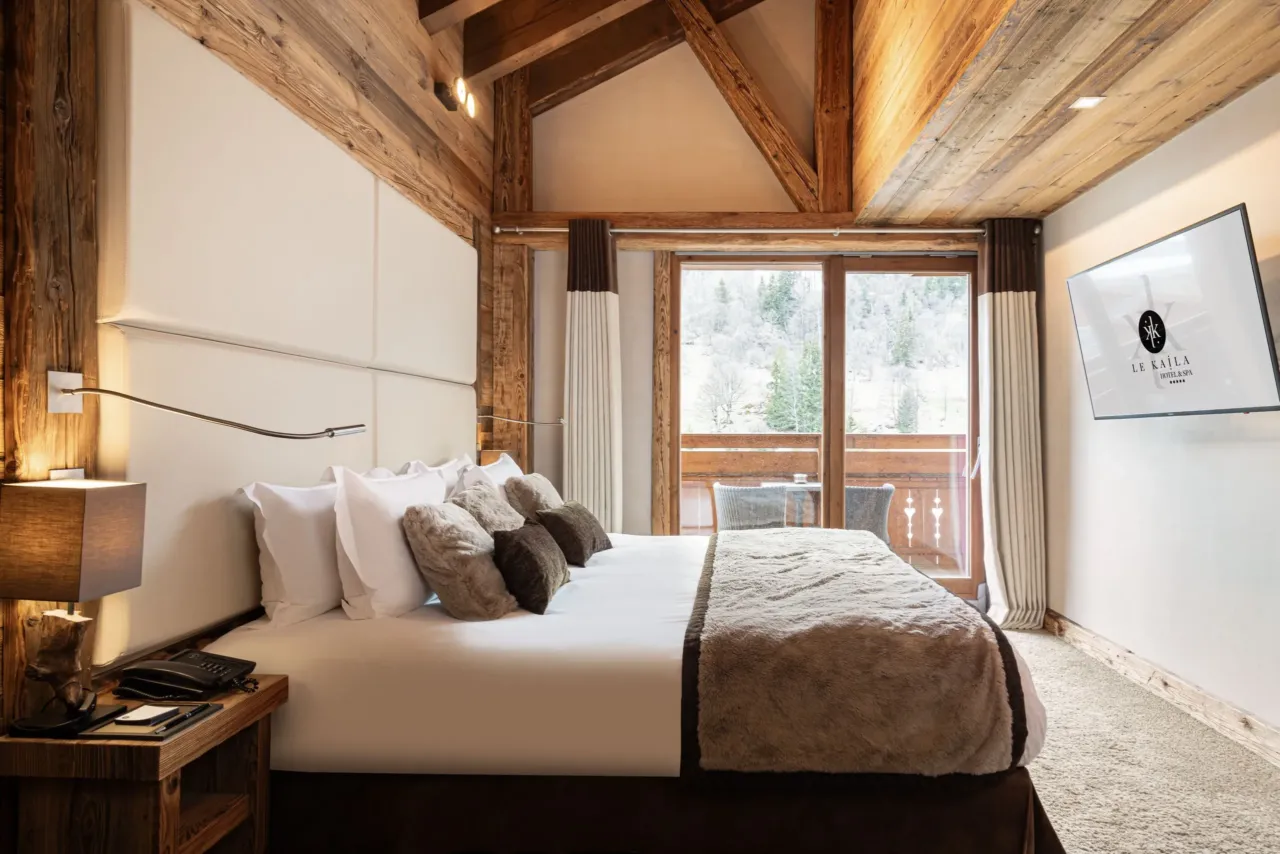 Chambre avec grand lit, tête de lit capitonnée et balcon donnant sur un versant boisé enneigé.